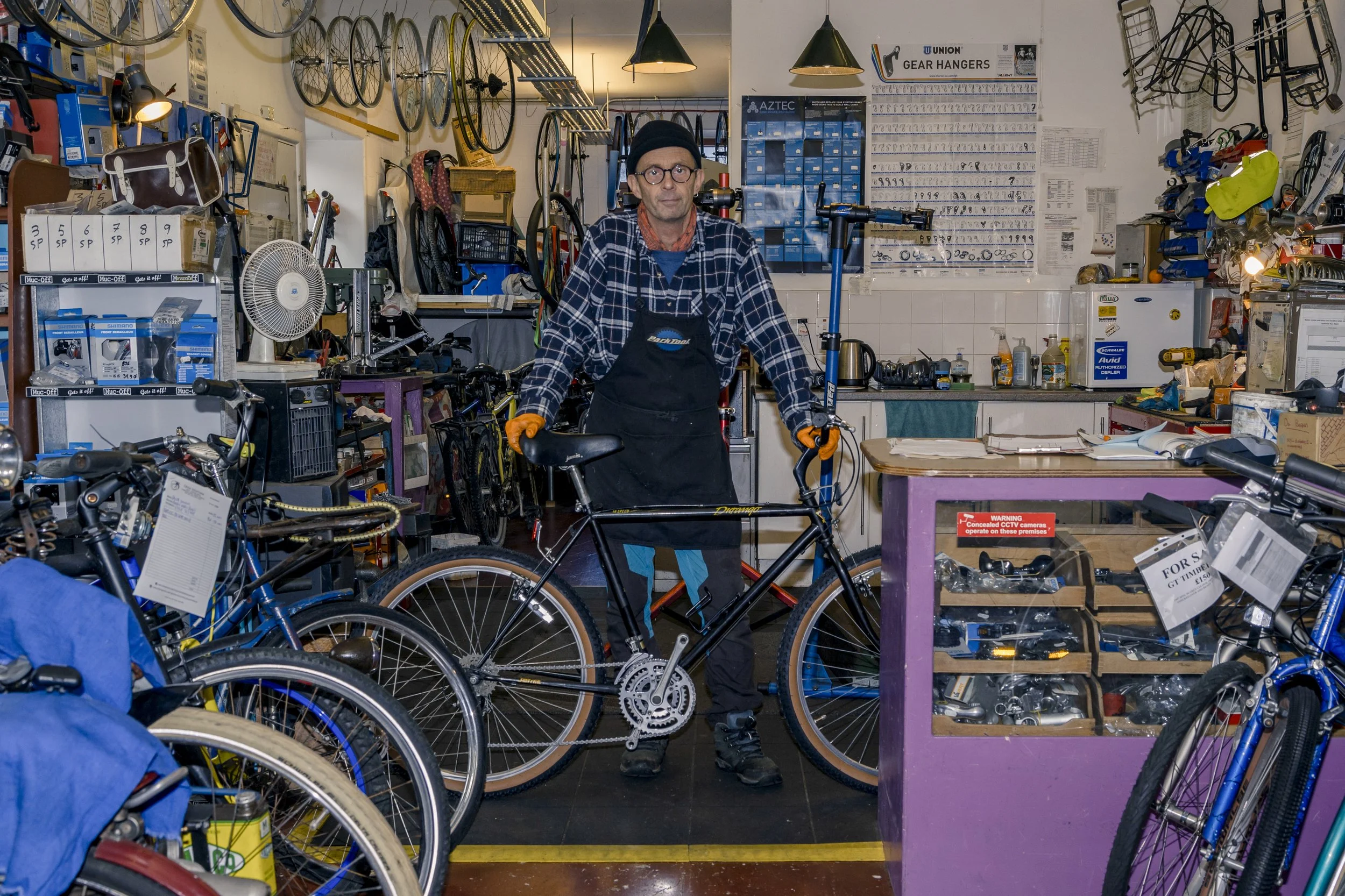 A man standing in a bicycle shop surrounded by various bicycles, bike parts, and tools. The man is wearing a black beanie, glasses, a plaid shirt, and gloves, holding a black bicycle frame.