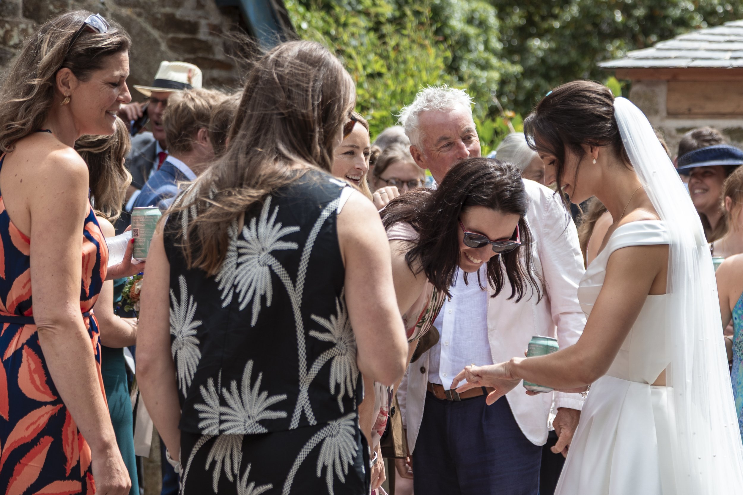 A bride in a white wedding dress and veil smiling and talking with a group of friends and family at a wedding outdoor reception.