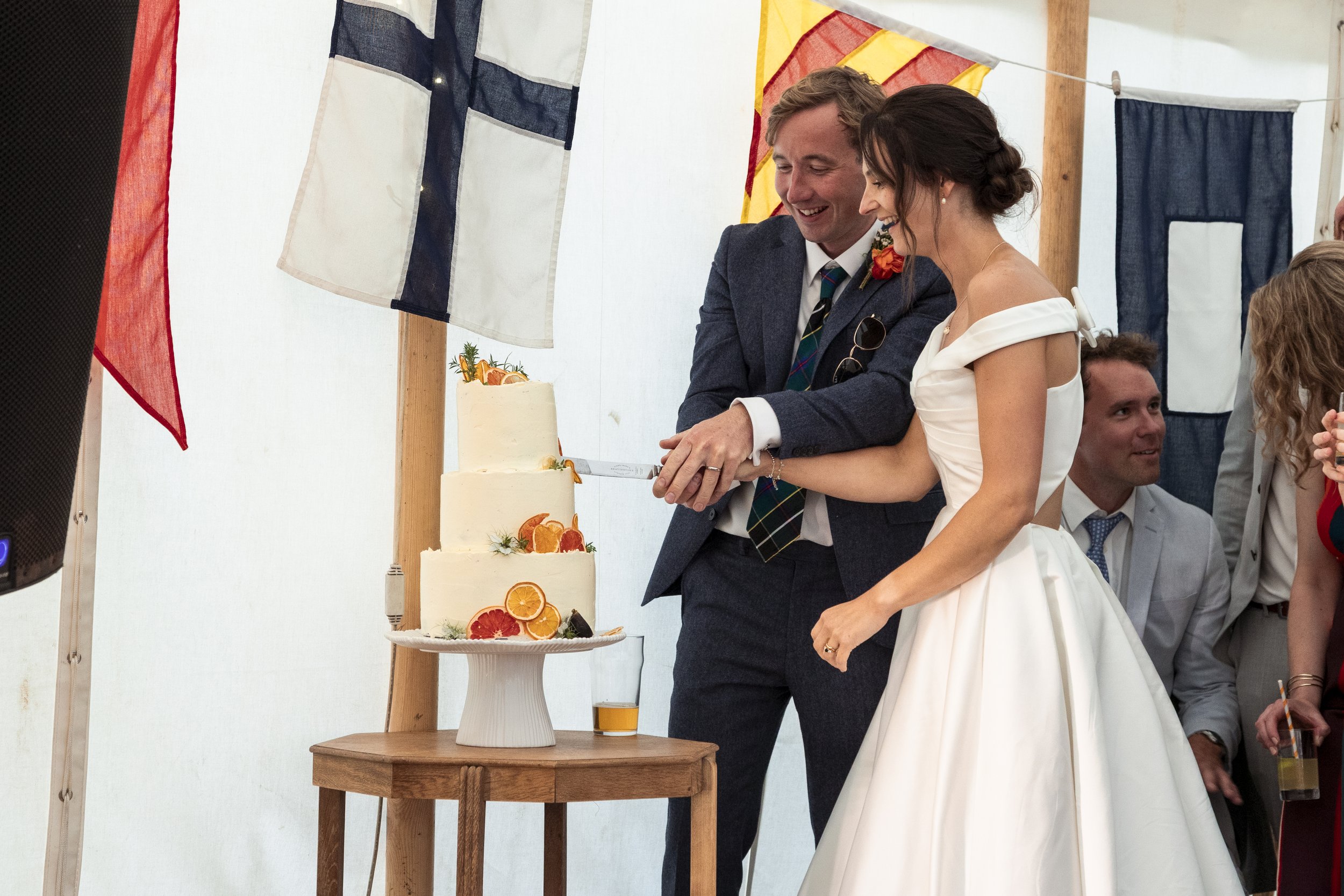 A bride and groom cutting a wedding cake decorated with fruit slices at a wedding celebration under Union Jack flags.