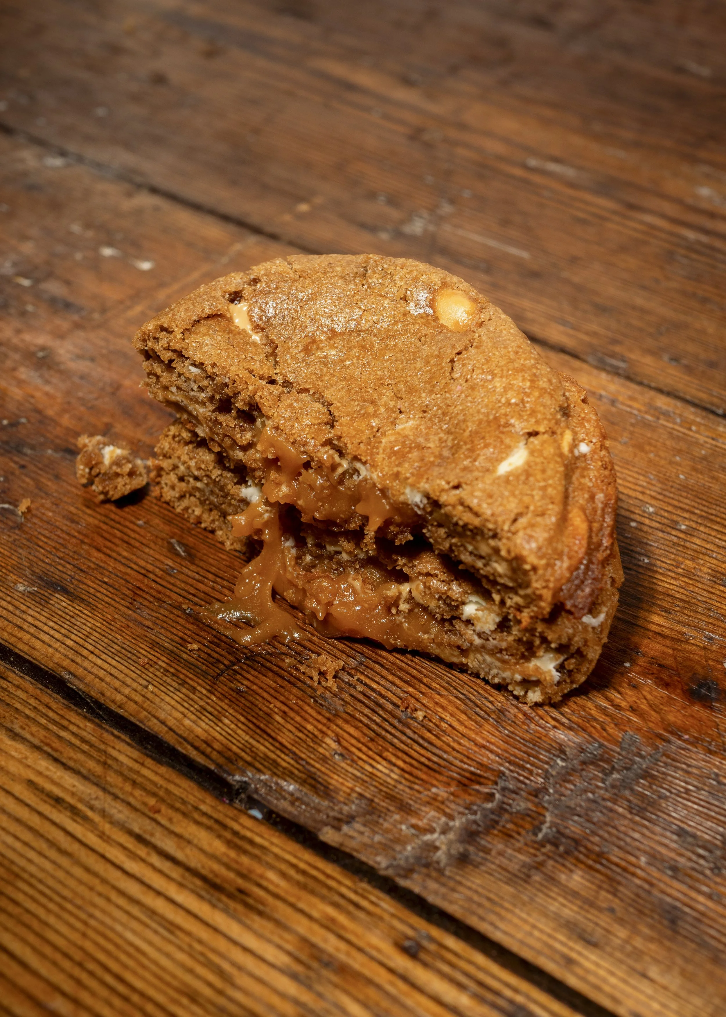 A partially eaten chocolate chip cookie with caramel filling on a wooden surface.