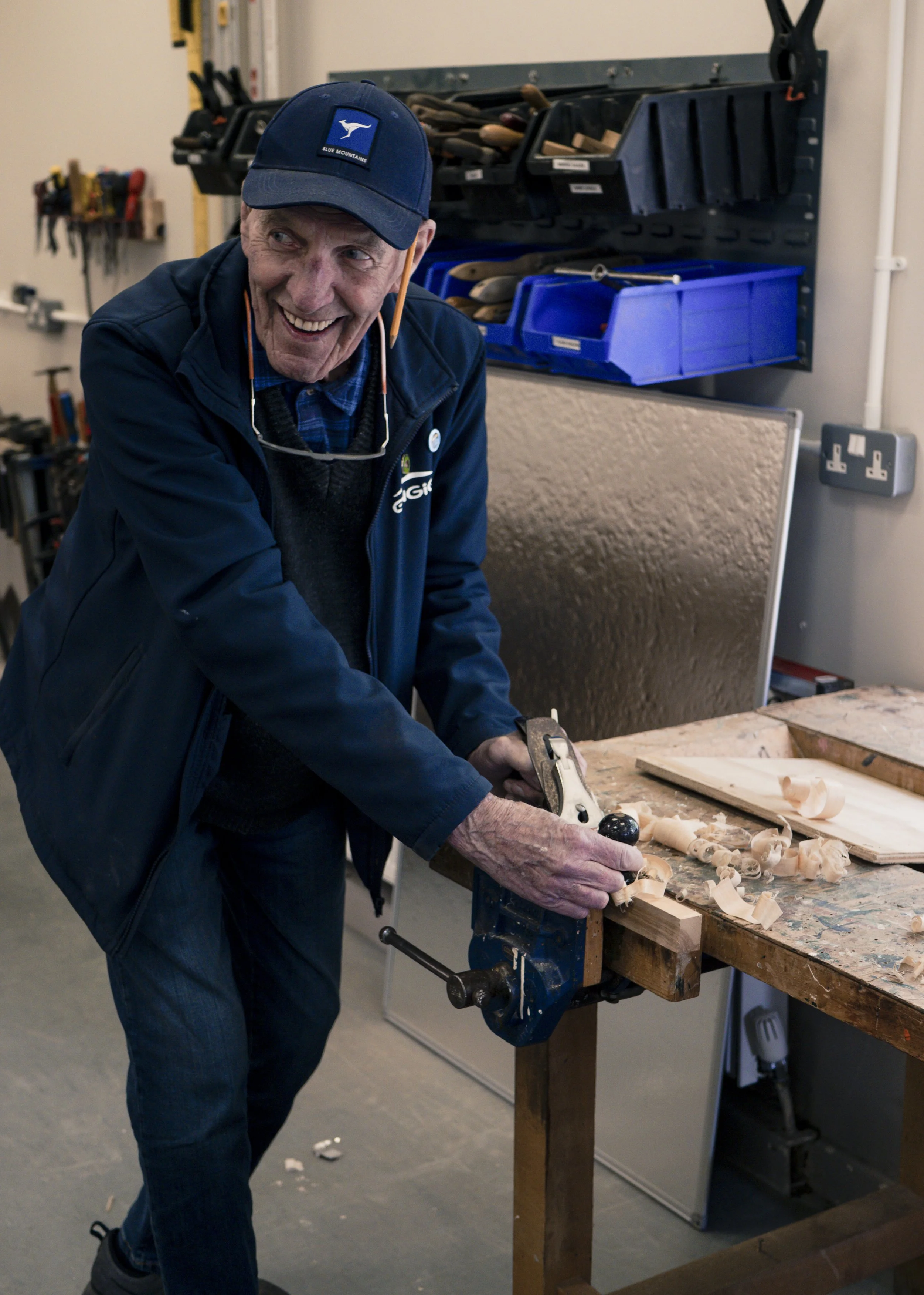 An elderly man wearing a blue cap, jacket, and plaid shirt is standing at a workbench carving wood with a hand plane.