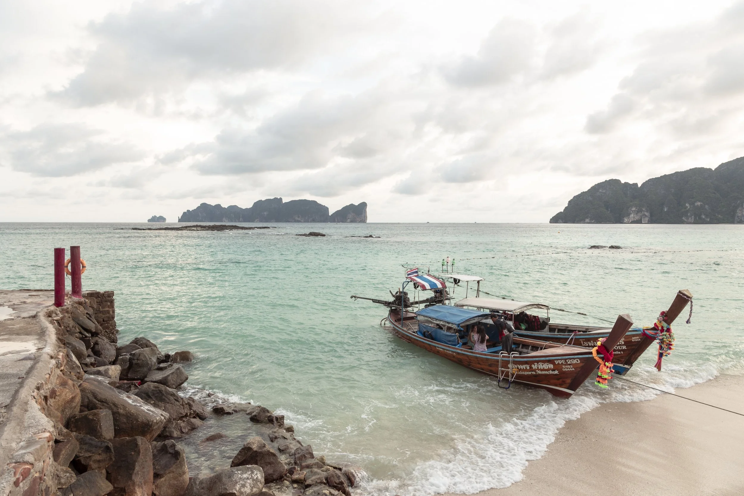 A boat docked at the sandy beach of a tropical island with limestone cliffs in the background under a cloudy sky.