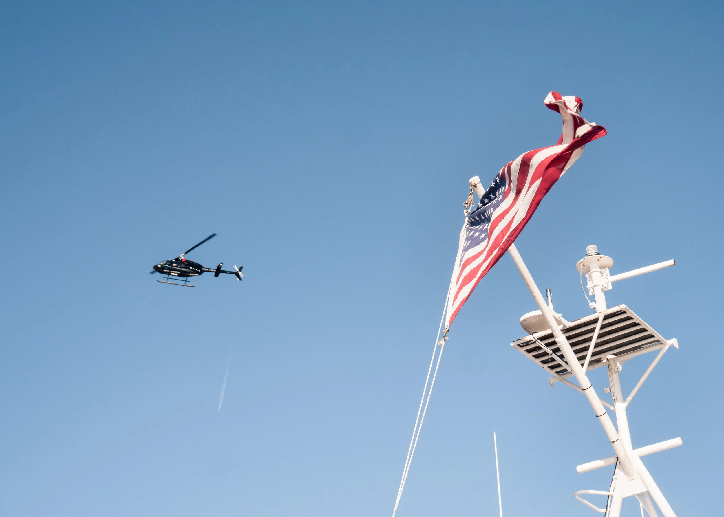 A helicopter flying in a clear blue sky near a boat's radar and an American flag.