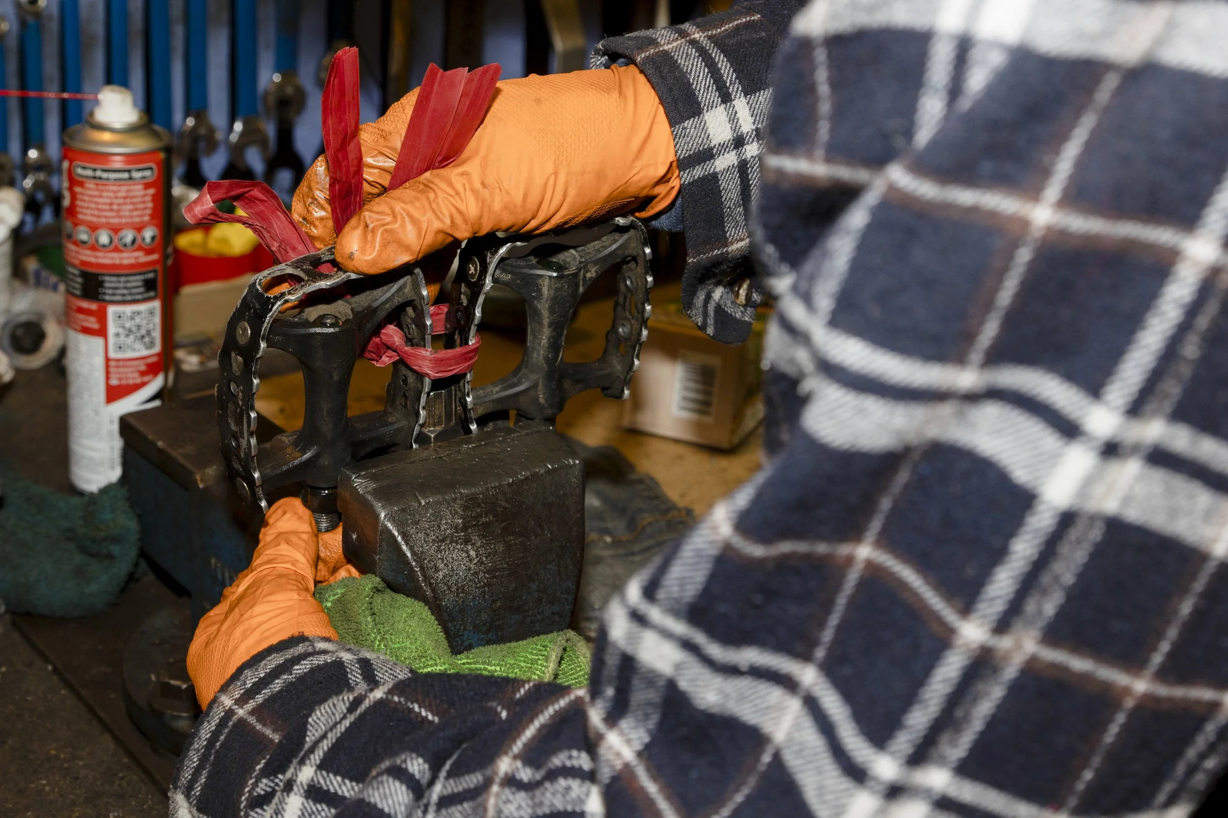 Person wearing plaid shirt and orange gloves working with a metal chain and black metal part in a workshop, with spray cans and tools in the background