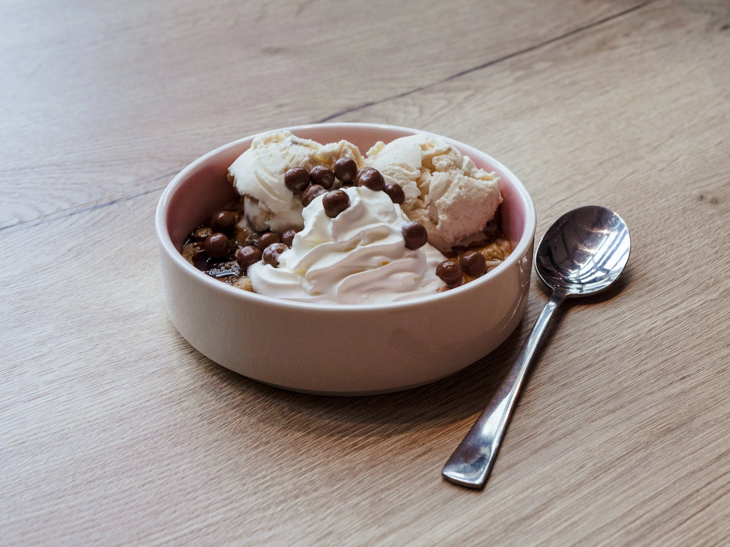 A bowl of ice cream topped with whipped cream, chocolate sprinkles, and chunks of vanilla ice cream, placed on a wooden table with a silver spoon beside it.