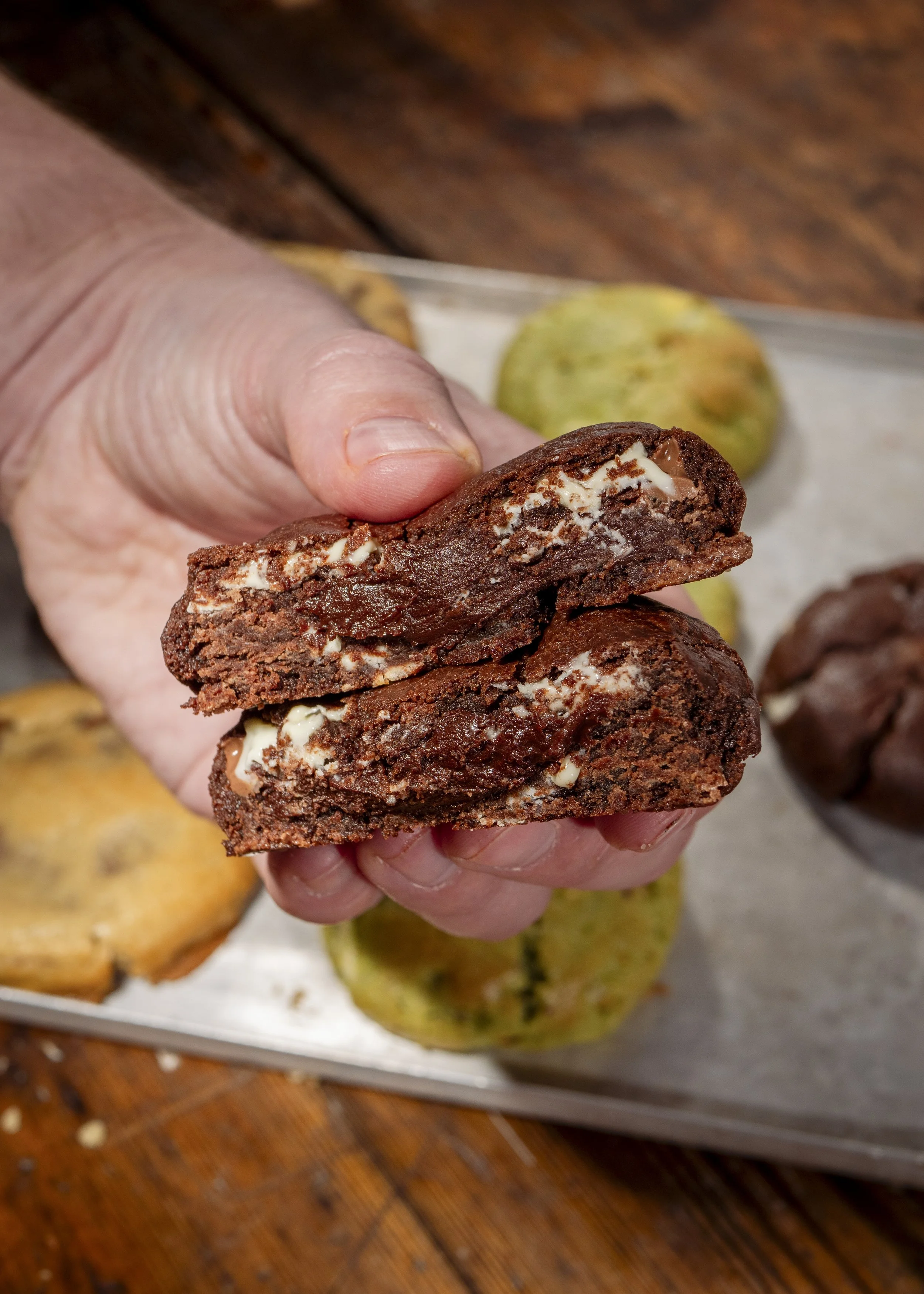 Close-up of a person's hand holding a broken chocolate cookie with white filling, with various cookies on a tray in the background.