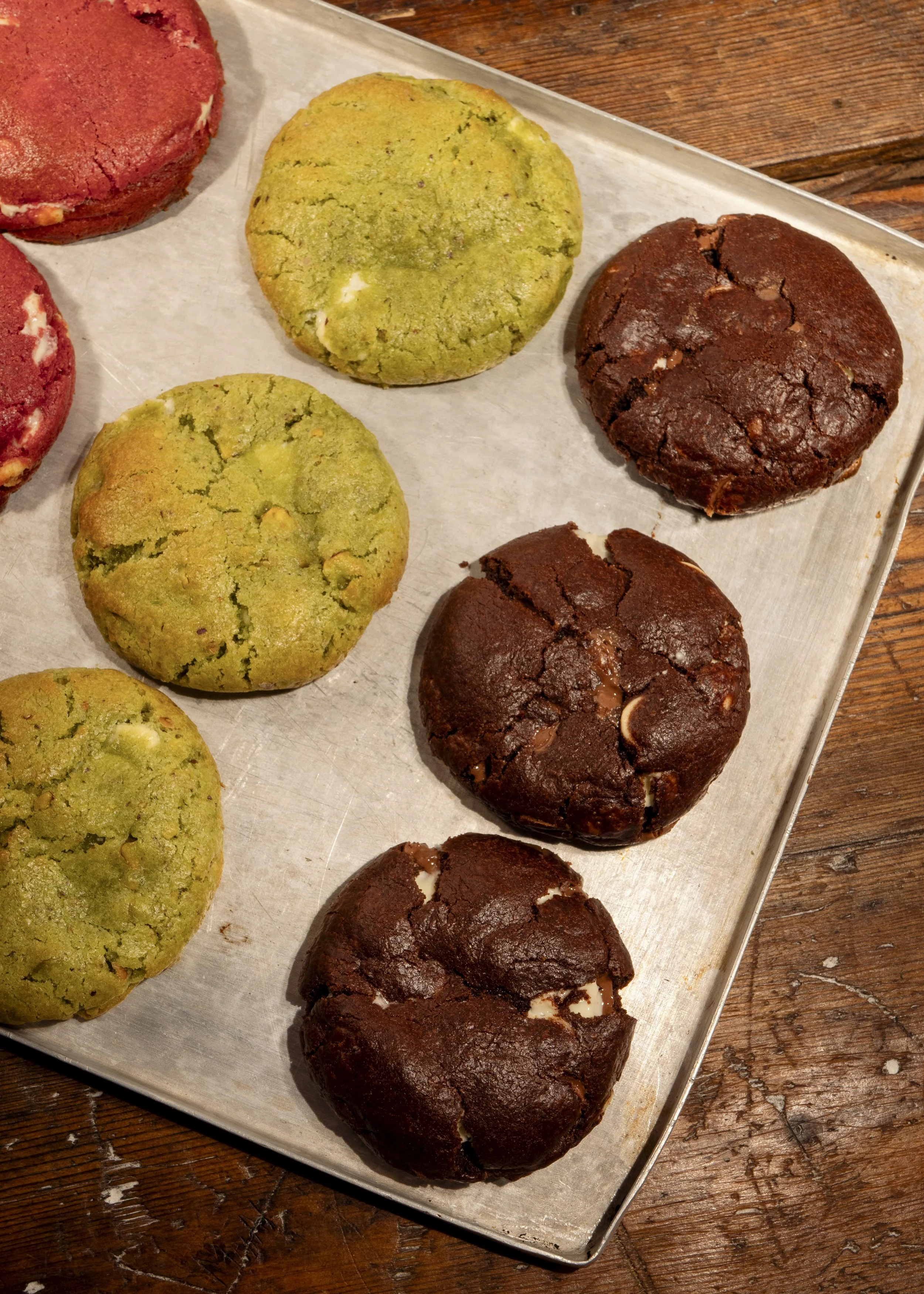 Six cookies on a baking sheet, with three green cookies and three chocolate cookies, placed on a wooden table.