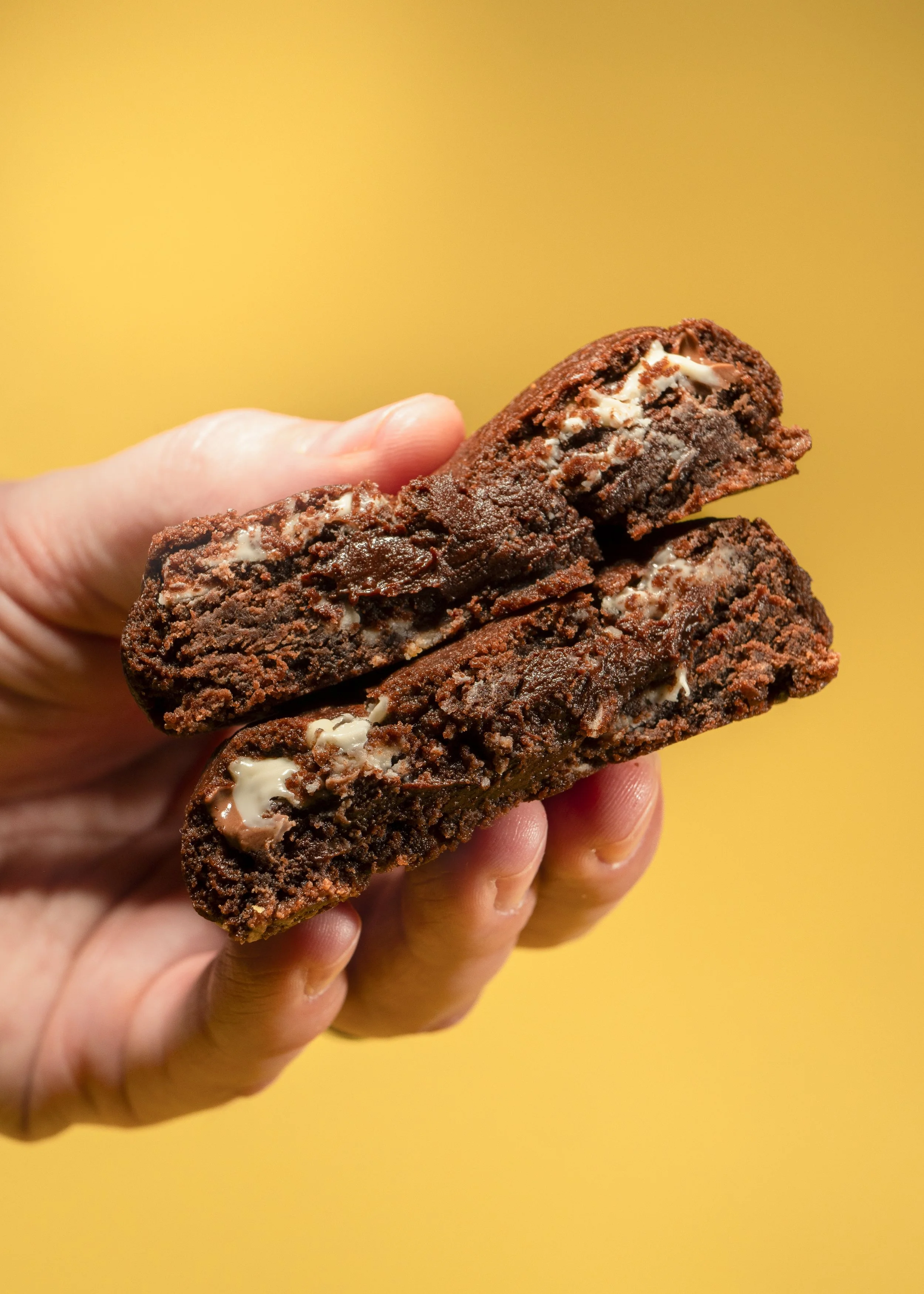 Close-up of a hand holding two chocolate cookies with white chocolate chunks against a yellow background.
