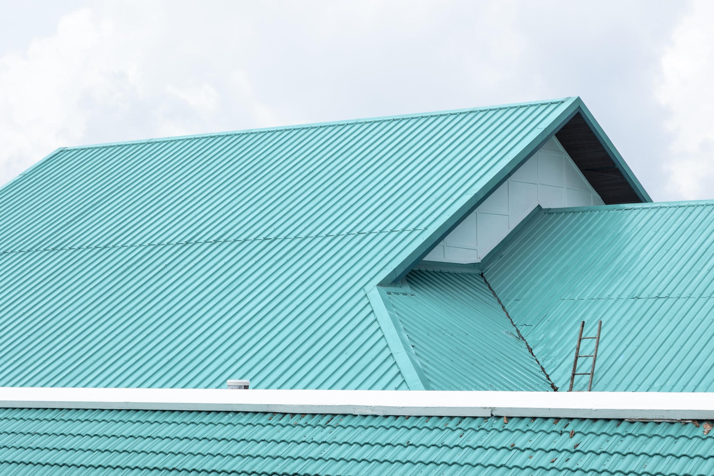 Close-up view of a teal corrugated metal roof with a small ladder leaning against it, and a cloudy sky in the background.