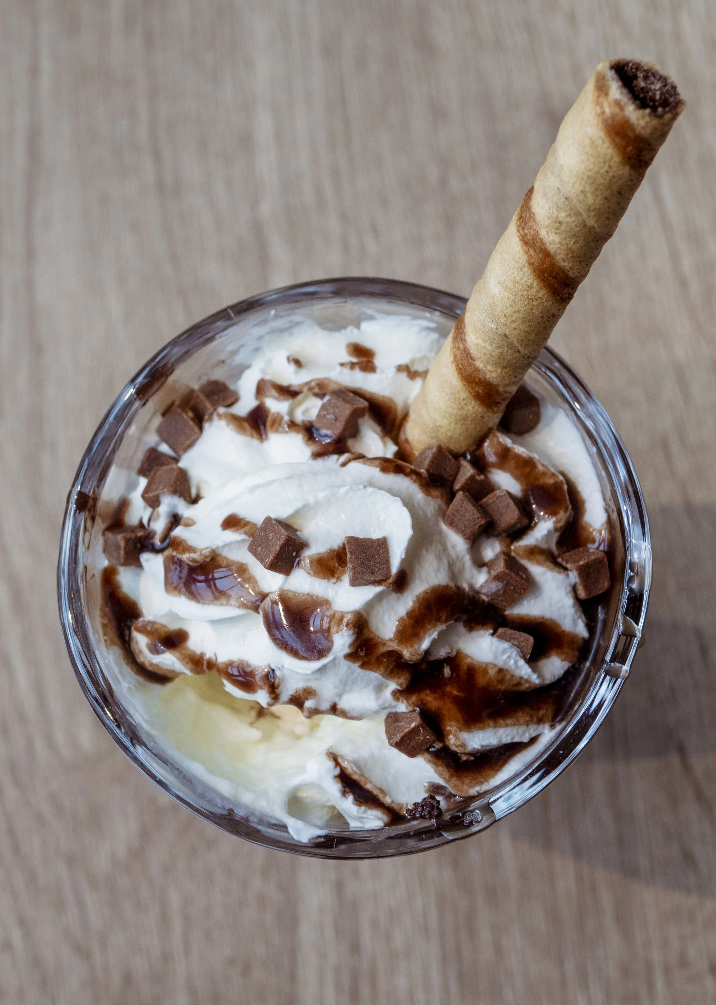 A glass of whipped cream topped with chocolate syrup, small chocolate chunks, and a rolled wafer cookie.