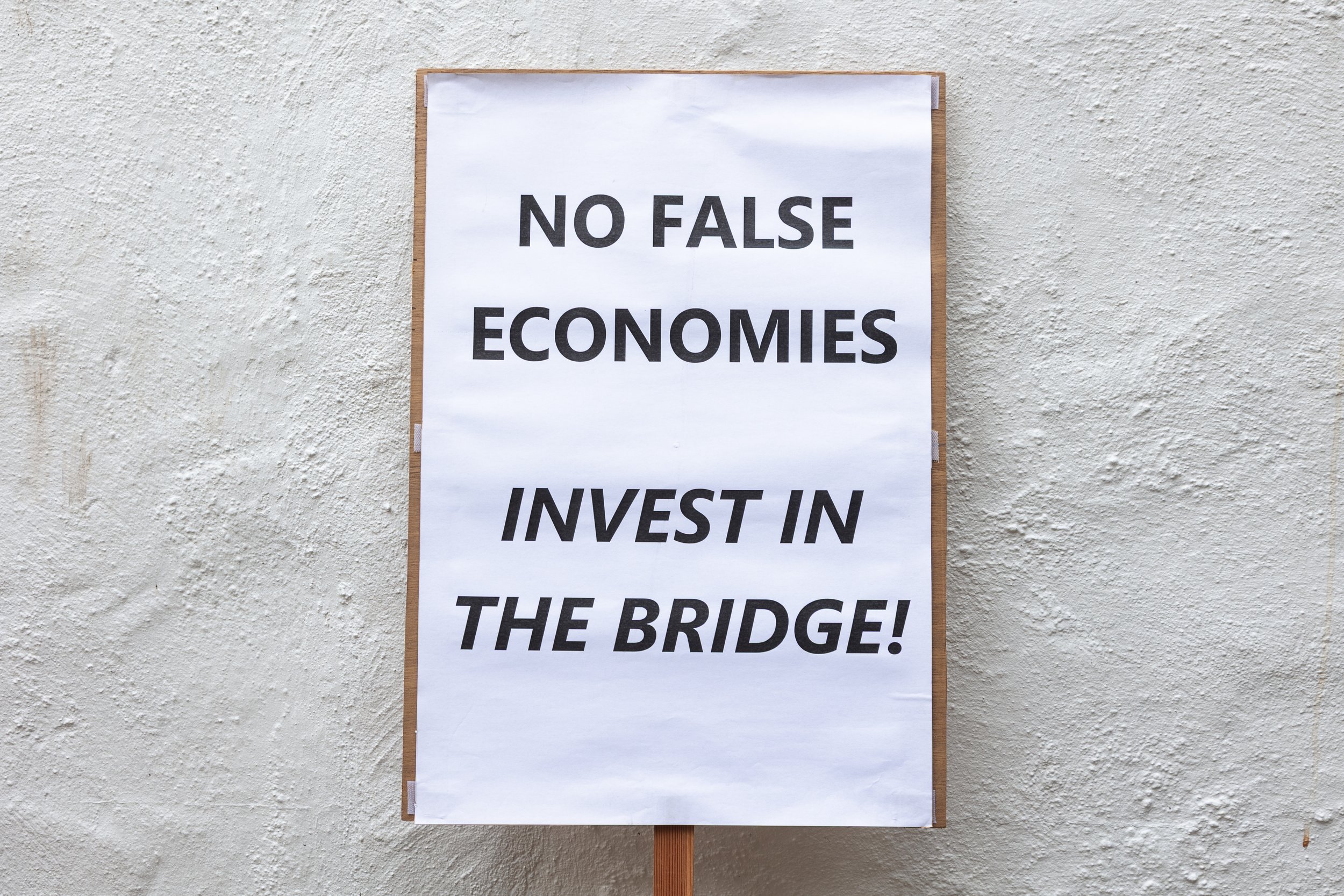 Sign with bold black text on white paper stating, 'NO FALSE ECONOMIES INVEST IN THE BRIDGE!', mounted on a wooden stand against a textured beige wall.