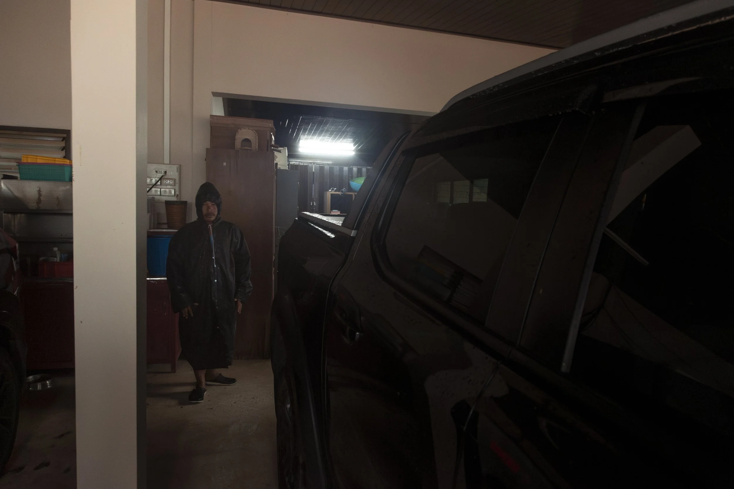 Person standing in a garage wearing a raincoat, with a black vehicle to the right and shelves with various items in the background.