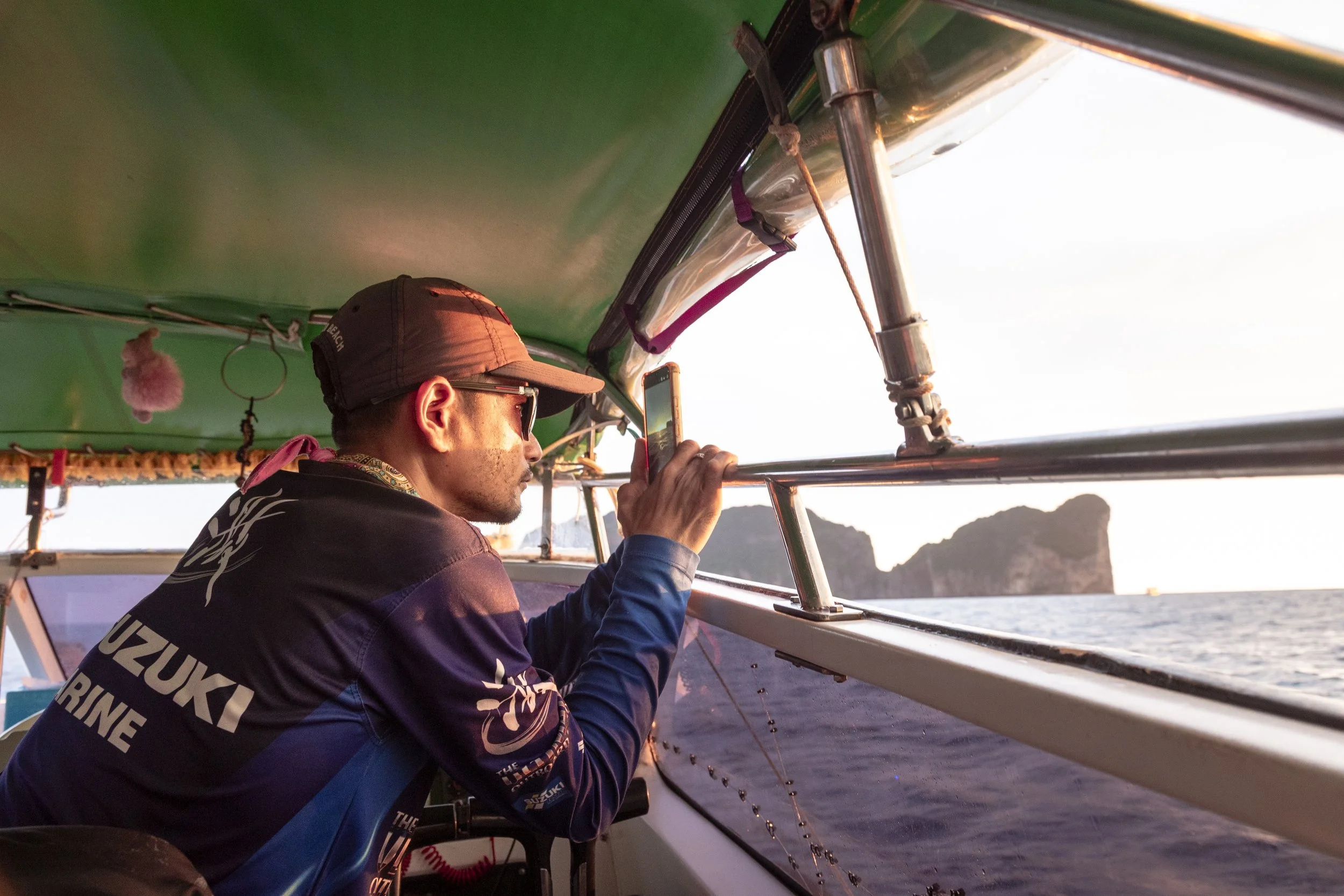 Man taking a photo or video with a smartphone on a boat during sunset, with a large rock formation visible in the water outside the boat.