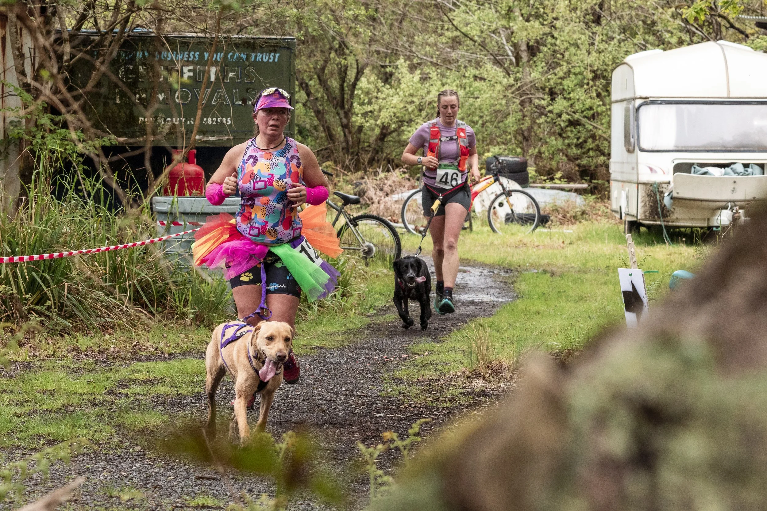 Two women participating in an outdoor event with dogs, one dressed in rainbow-colored clothing and a tutu, the other in athletic wear, running along a wooded trail with their dogs.