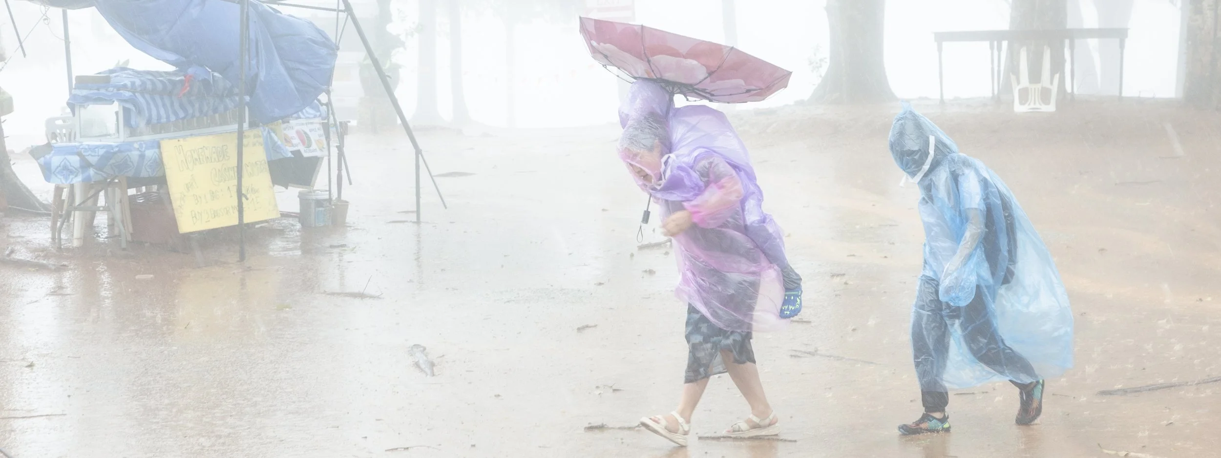 Two children wearing raincoats and sandals walk in heavy rain, holding umbrellas in a foggy outdoor setting with a vendor stand nearby.
