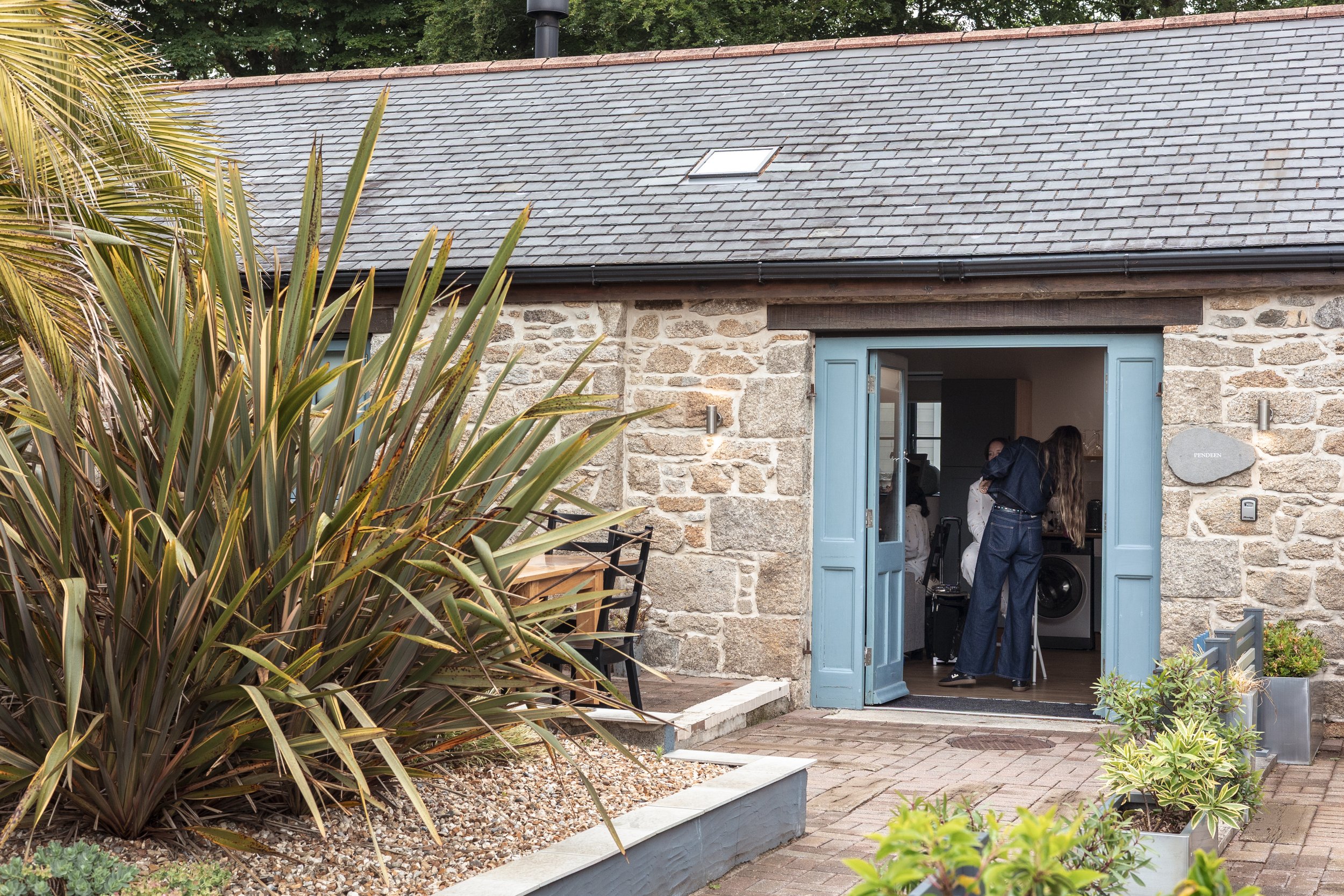 View of a house with a stone exterior and light blue door open to reveal people inside. The scene includes a colorful garden with large, spiky plants in the foreground and potted plants on the right.