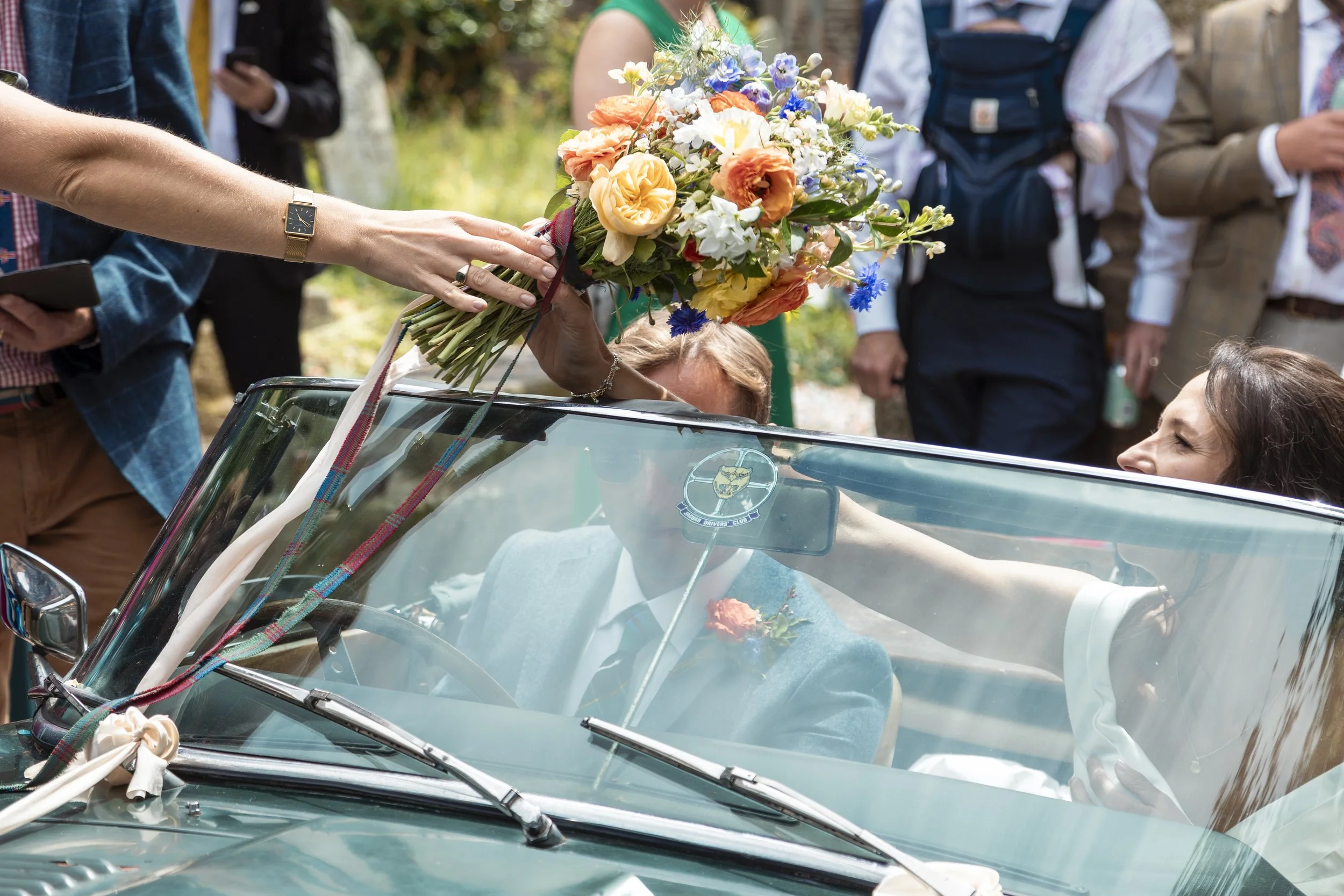 A woman sitting in an open-top vintage car receiving a bouquet of flowers from a person outside the car, with several people standing around and watching.
