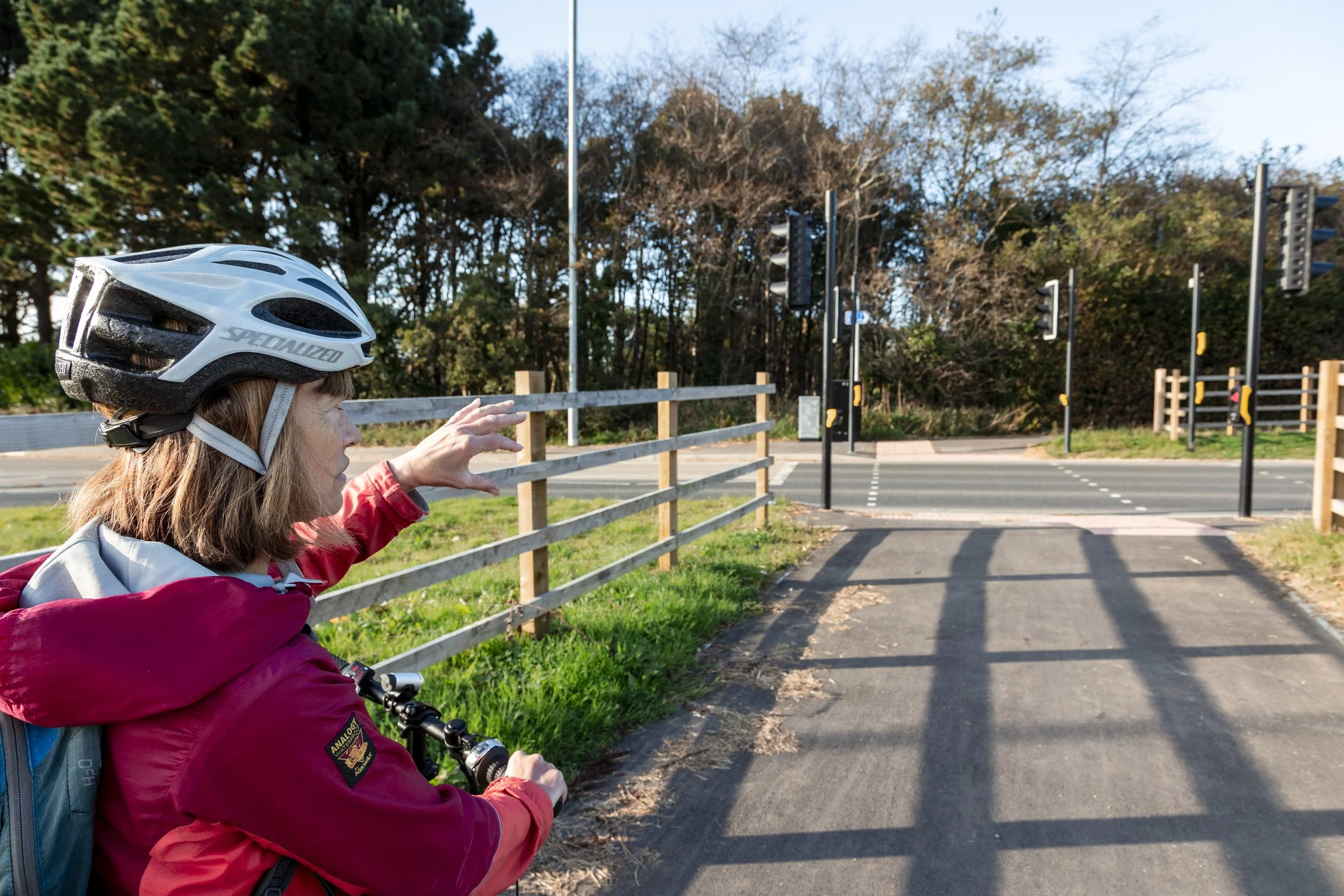 A woman in a red jacket and helmet points towards a pedestrian crossing with traffic lights at a bicycle path intersection.