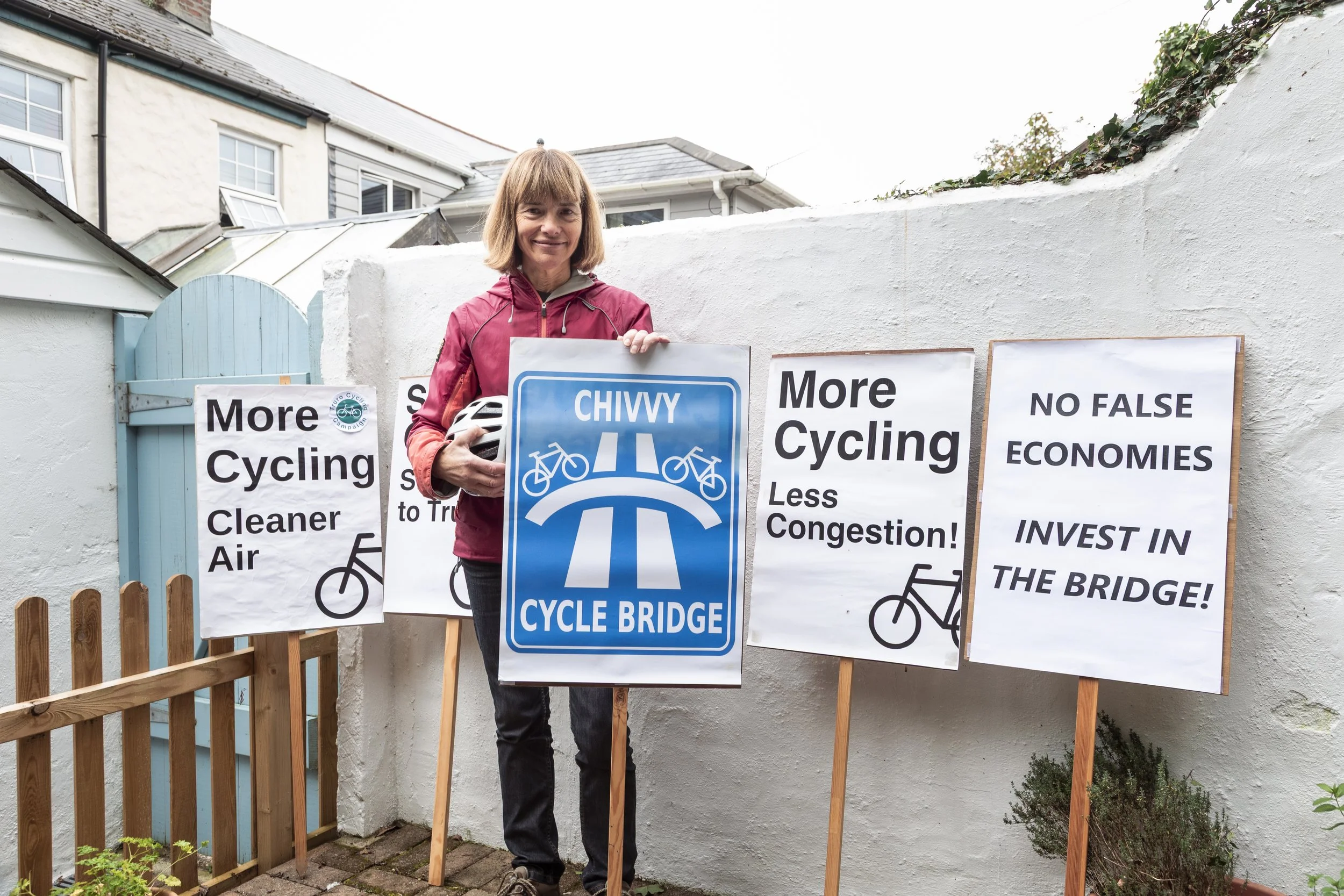 Woman holding a bicycle helmet standing next to protest signs advocating for more cycling infrastructure and cleaner air outside a white wall with houses in the background.
