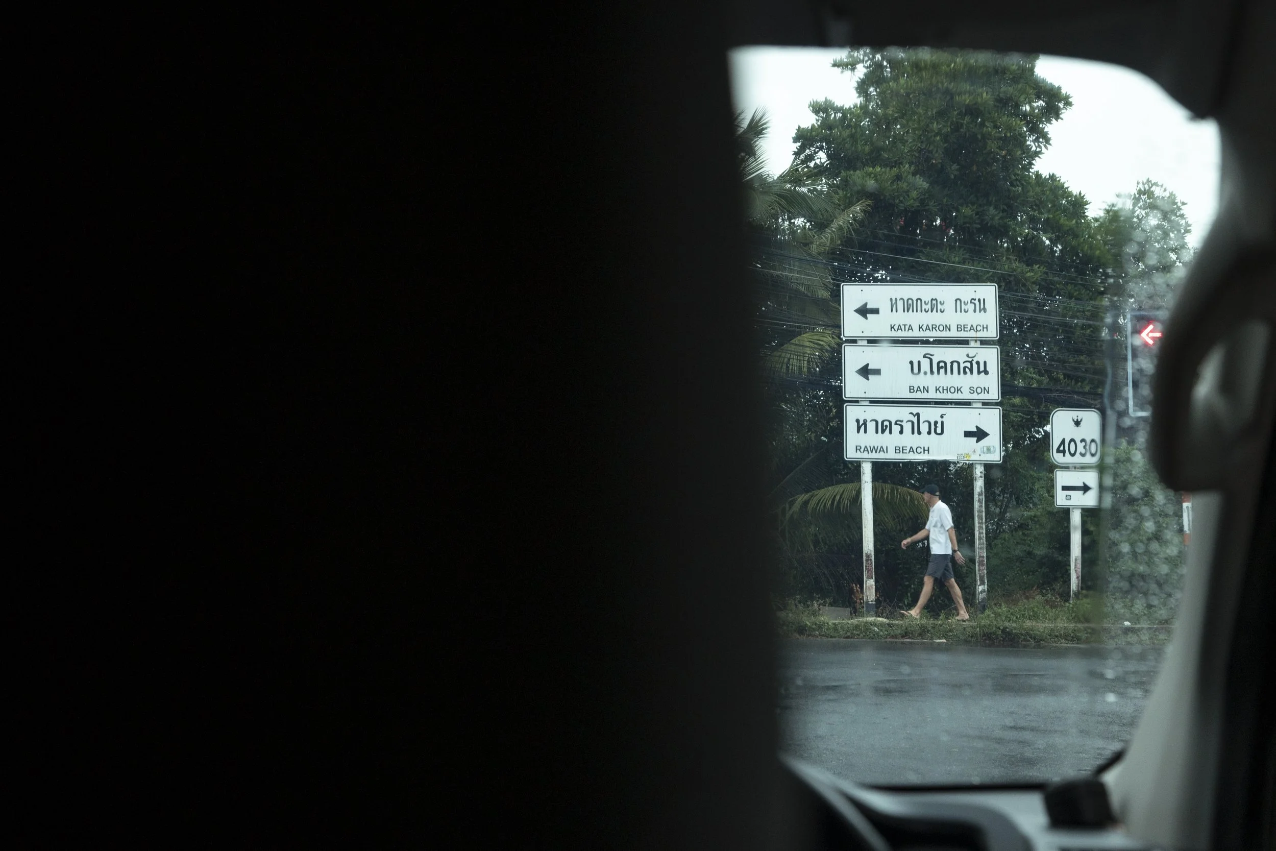 View through a car window showing directional road signs pointing to Kata Karon Beach, Ban Khok Son, and Rawai Beach, with a person walking on the sidewalk and rainy weather.
