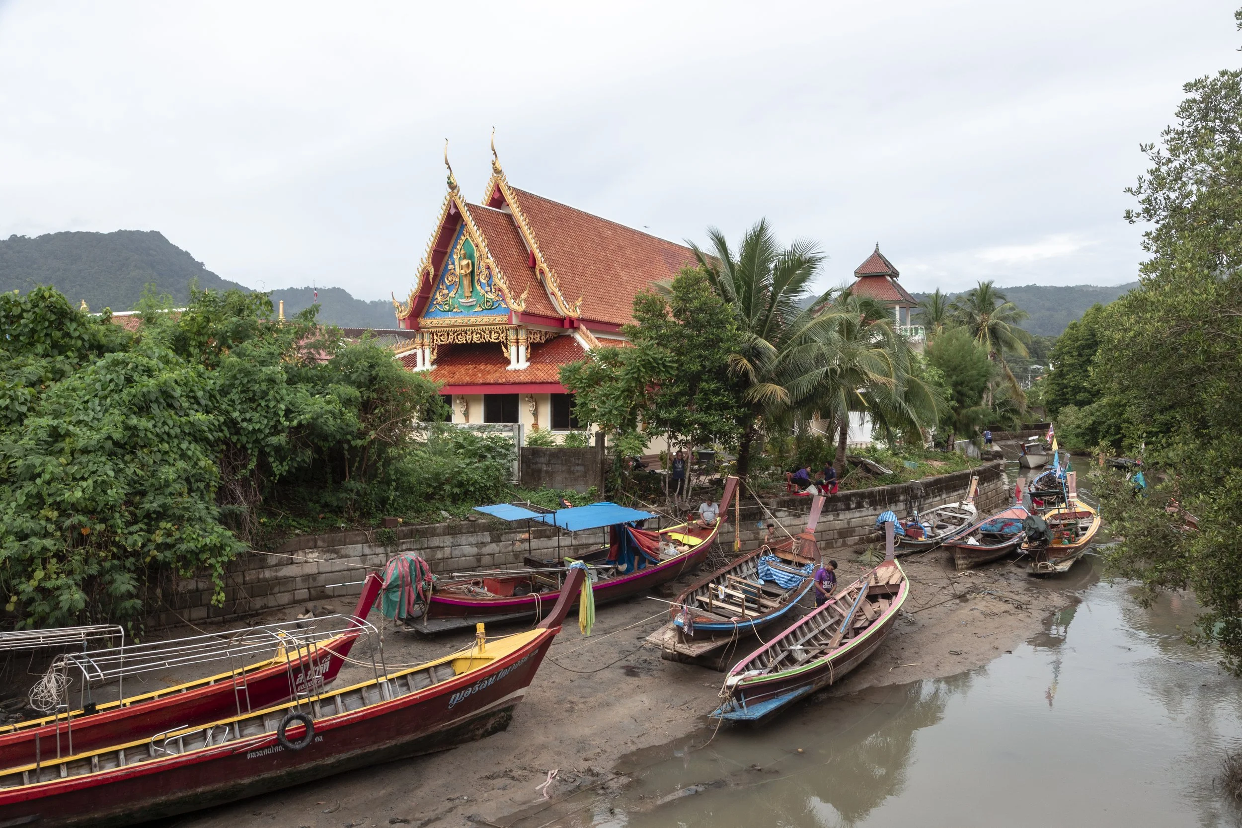 Traditional Thai temple with ornate roof decorations near a river, flanked by trees and boats docked along the riverbank.