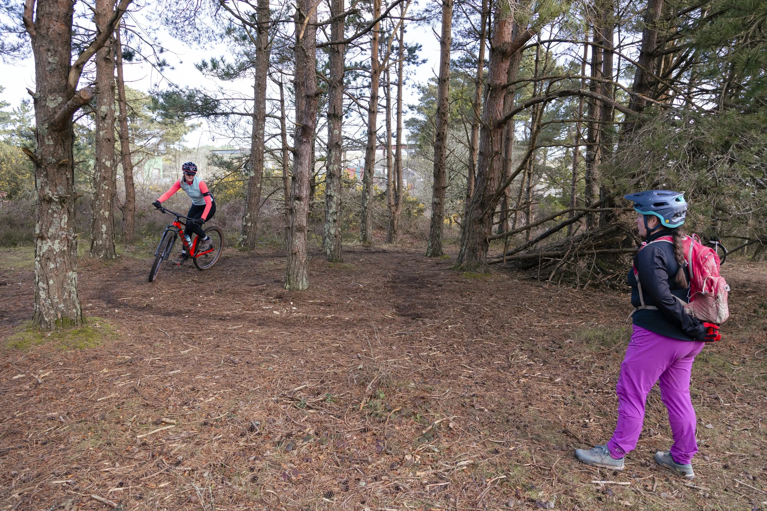 Two women in outdoor gear with helmets and backpacks in a wooded area; one woman is riding a mountain bike, while the other stands on the trail watching.
