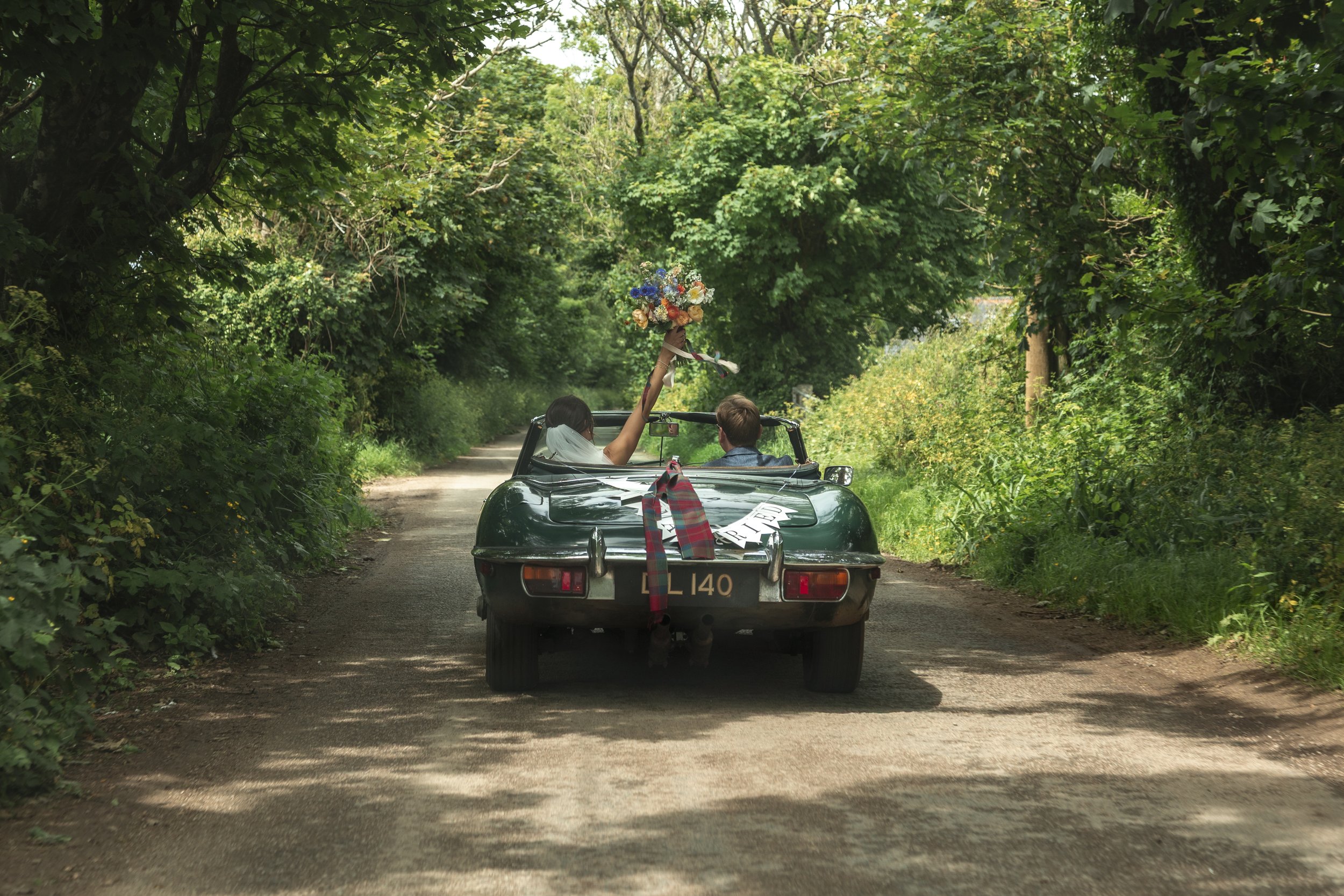 A green convertible car driving on a dirt road surrounded by lush green trees, with a bride and groom inside. The bride is holding a bouquet of flowers raised up, and the groom is sitting next to her. The car has signs on the back, including a red an