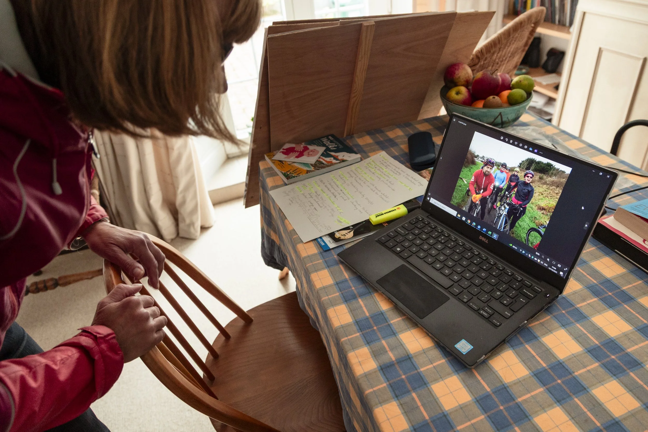 A woman leaning over a wooden chair to look at a laptop on a dining table. The laptop screen displays a photo of five cyclists outdoors. The table has a bowl of assorted apples, a few magazines, a set of papers with notes, a highlighter, and a remote control. The setup is on a checkered tablecloth.