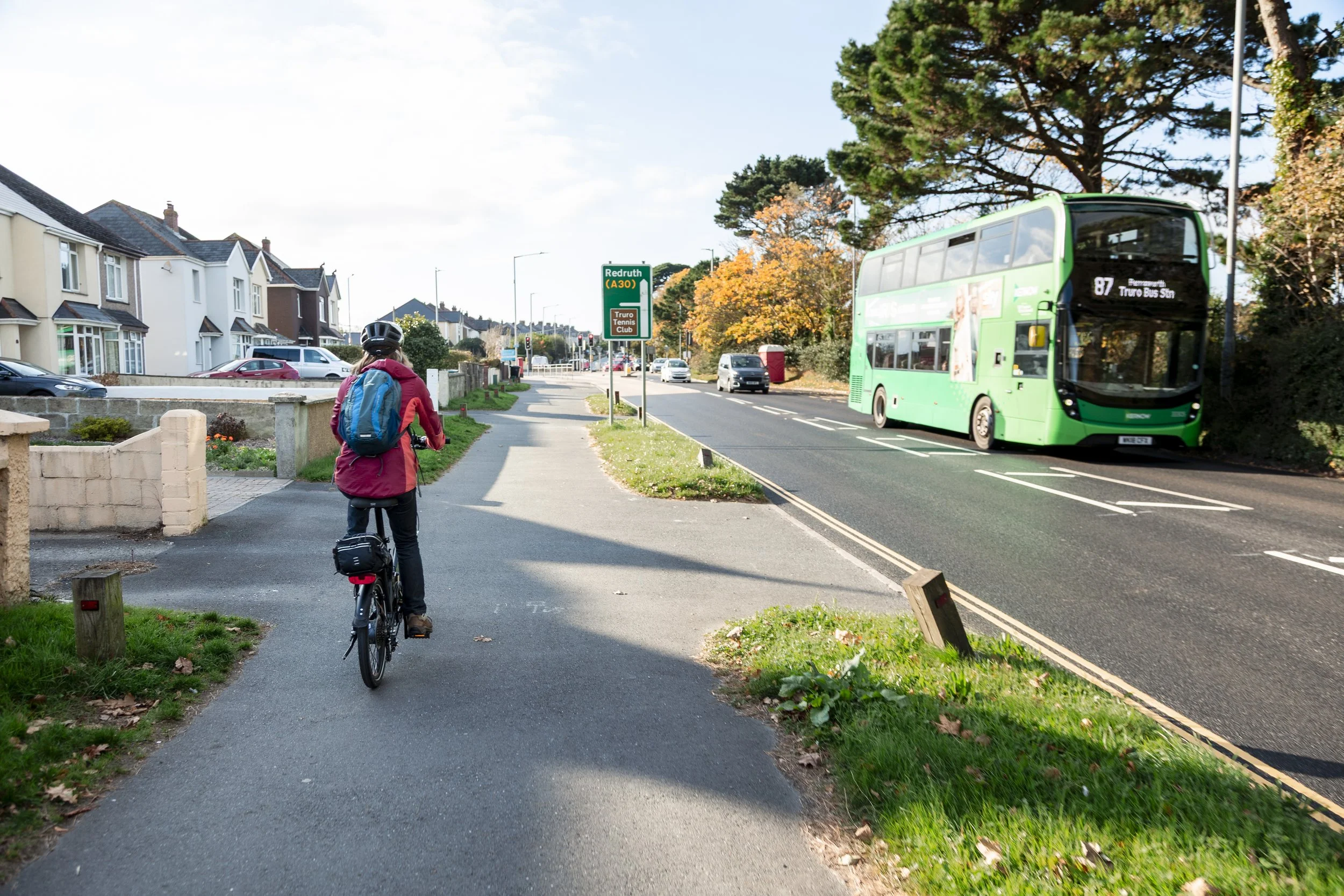 A person riding a bicycle on a sidewalk next to a street with a green bus in the background, residential houses on the left, and trees with autumn leaves.