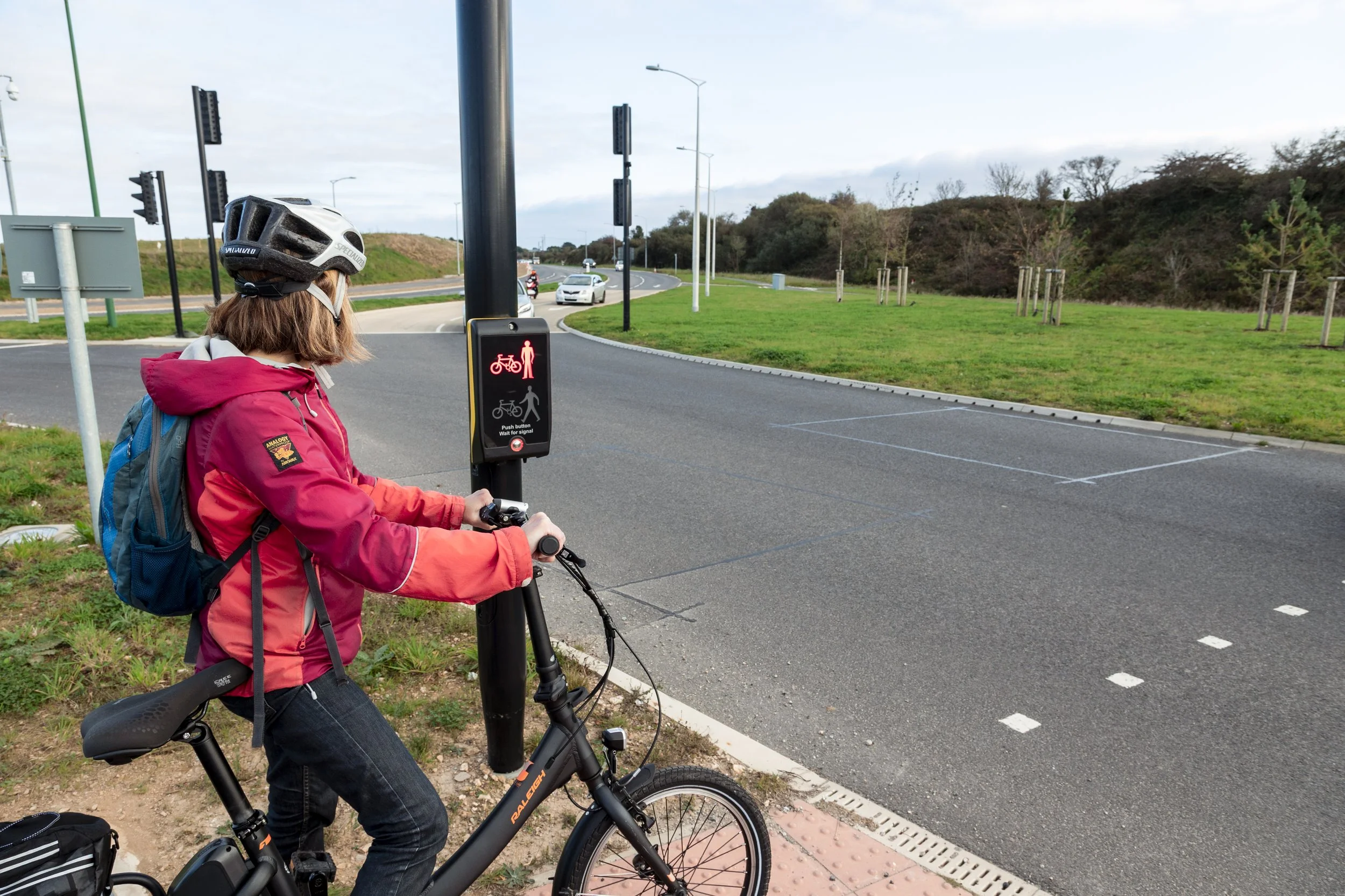 A woman wearing a red jacket and a helmet, with a backpack, on her bicycle at a pedestrian and bicycle signal crossing a street.