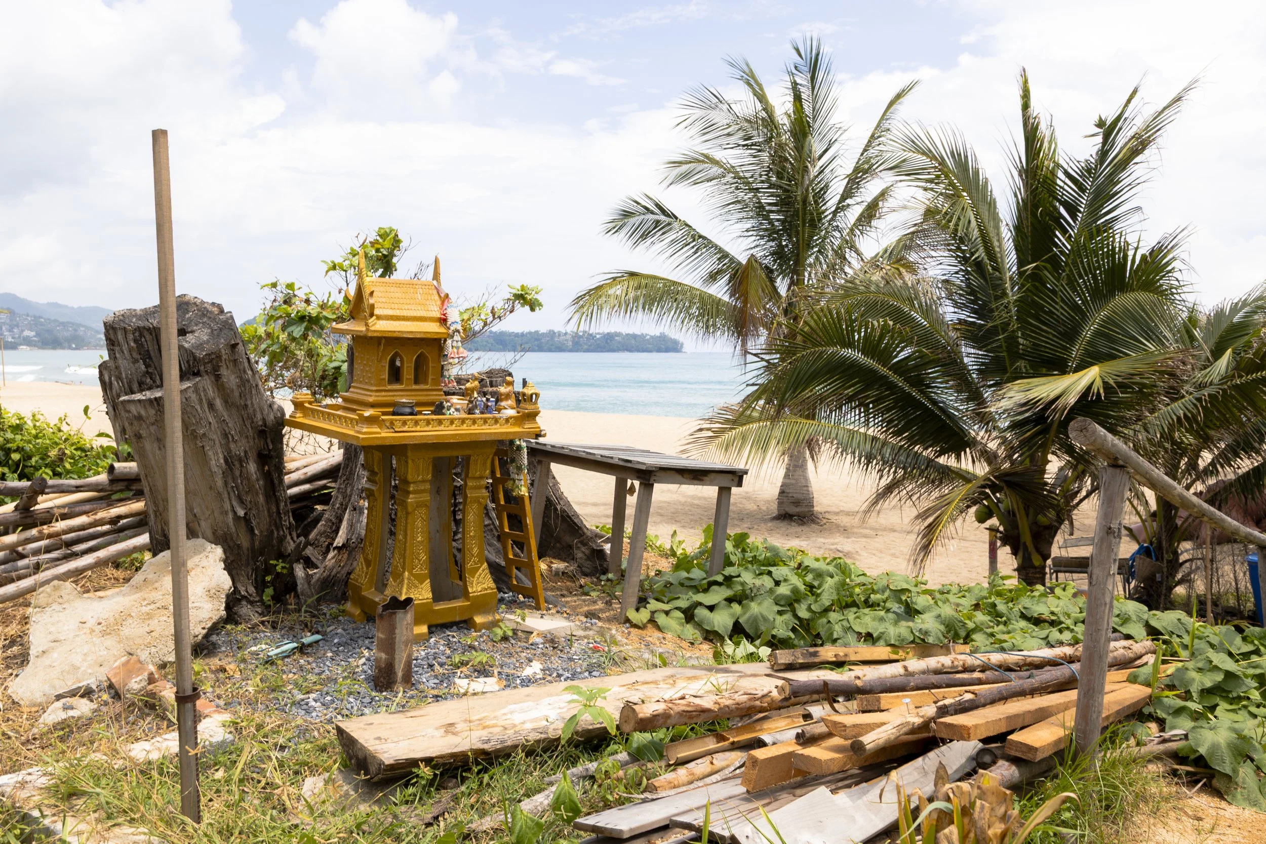 A small shrine on a sandy beach in Phuket, Thailand, with palm trees, a body of water, and a partly cloudy sky in the background.
