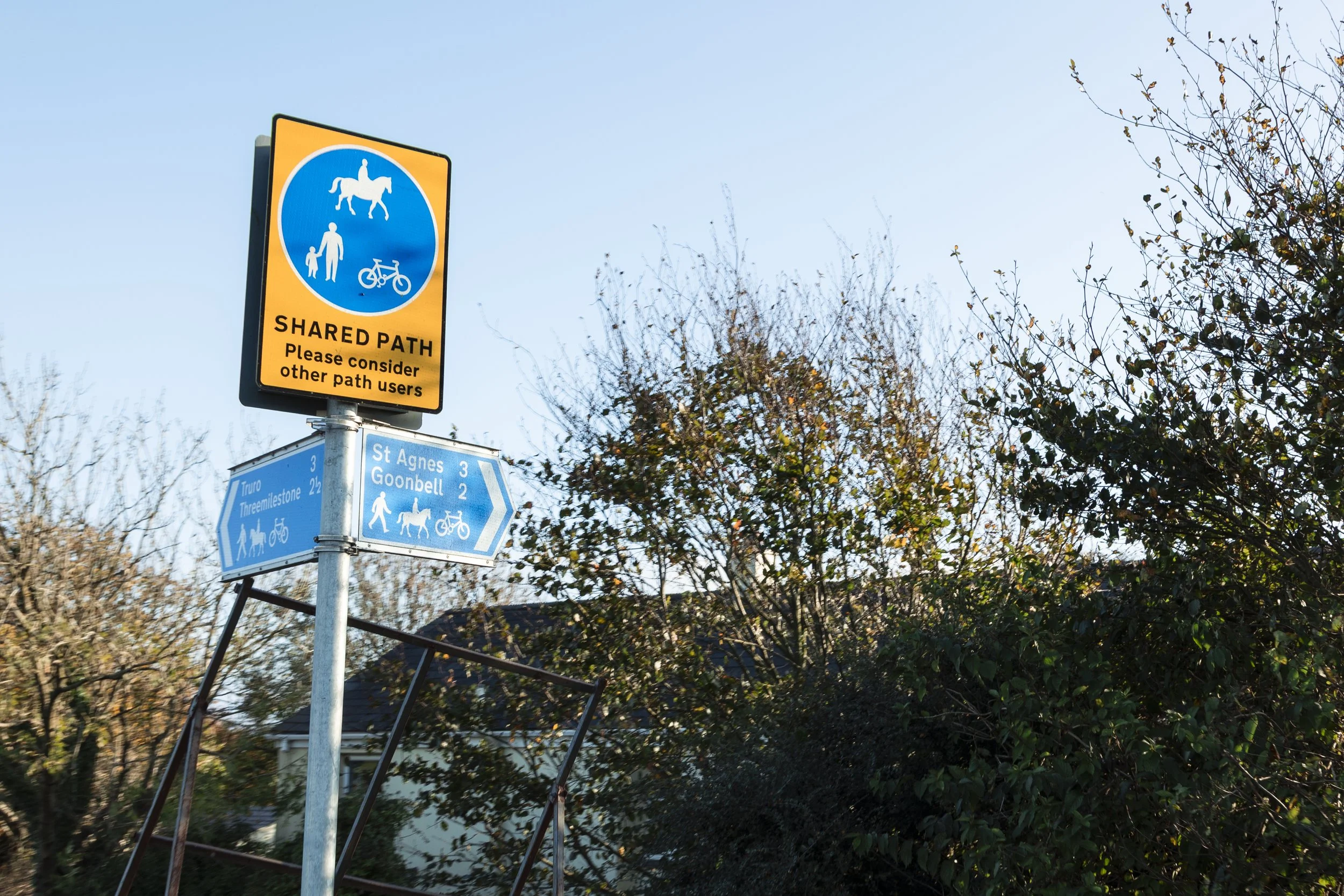 A signpost with a yellow and black shared path sign for pedestrians, cyclists, and horse riders, with a note to consider other users. Below it are blue directional signs indicating distances to Truro, Threemilestone, St Agnes, and Goonbell along a sh