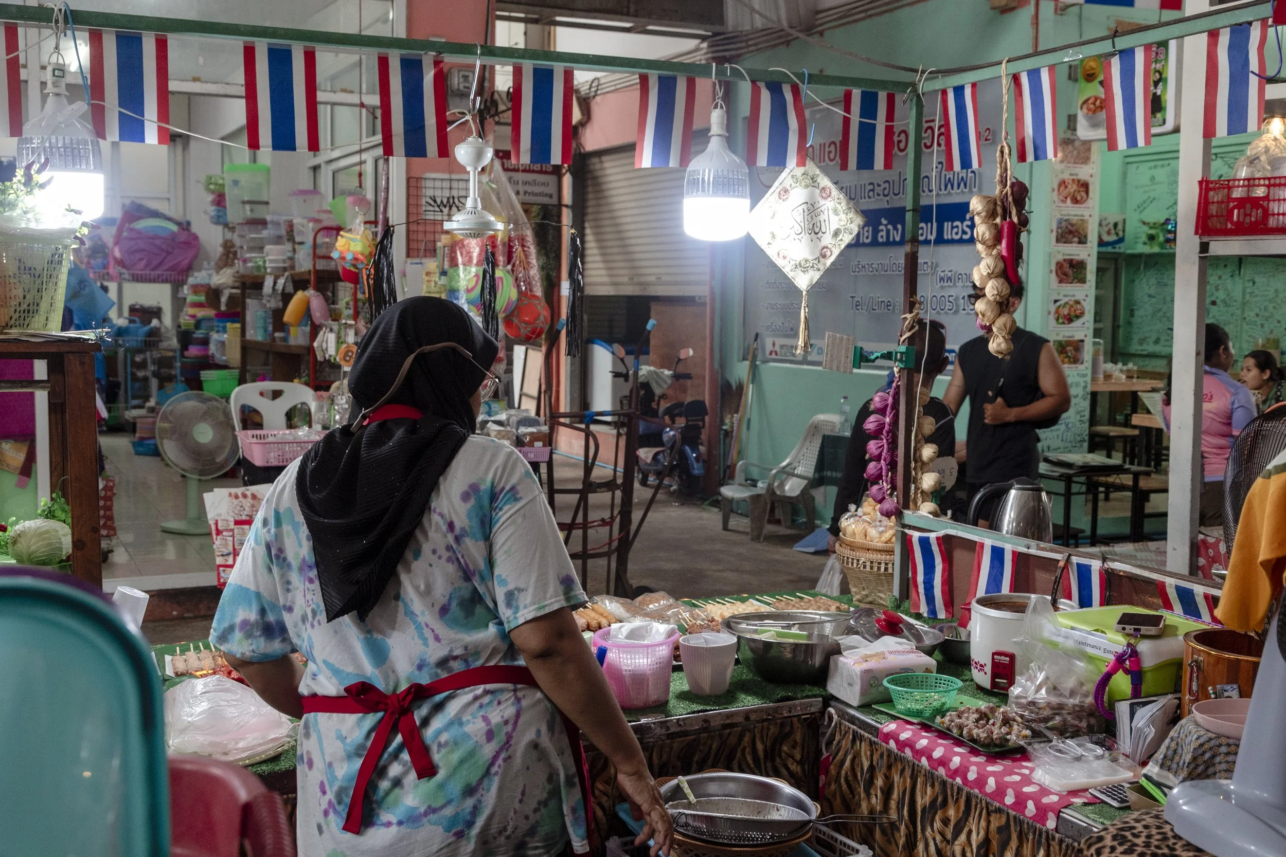 A woman wearing a black headscarf and a floral shirt working at a street stall selling food items at night, with colorful decorations and other vendors around.
