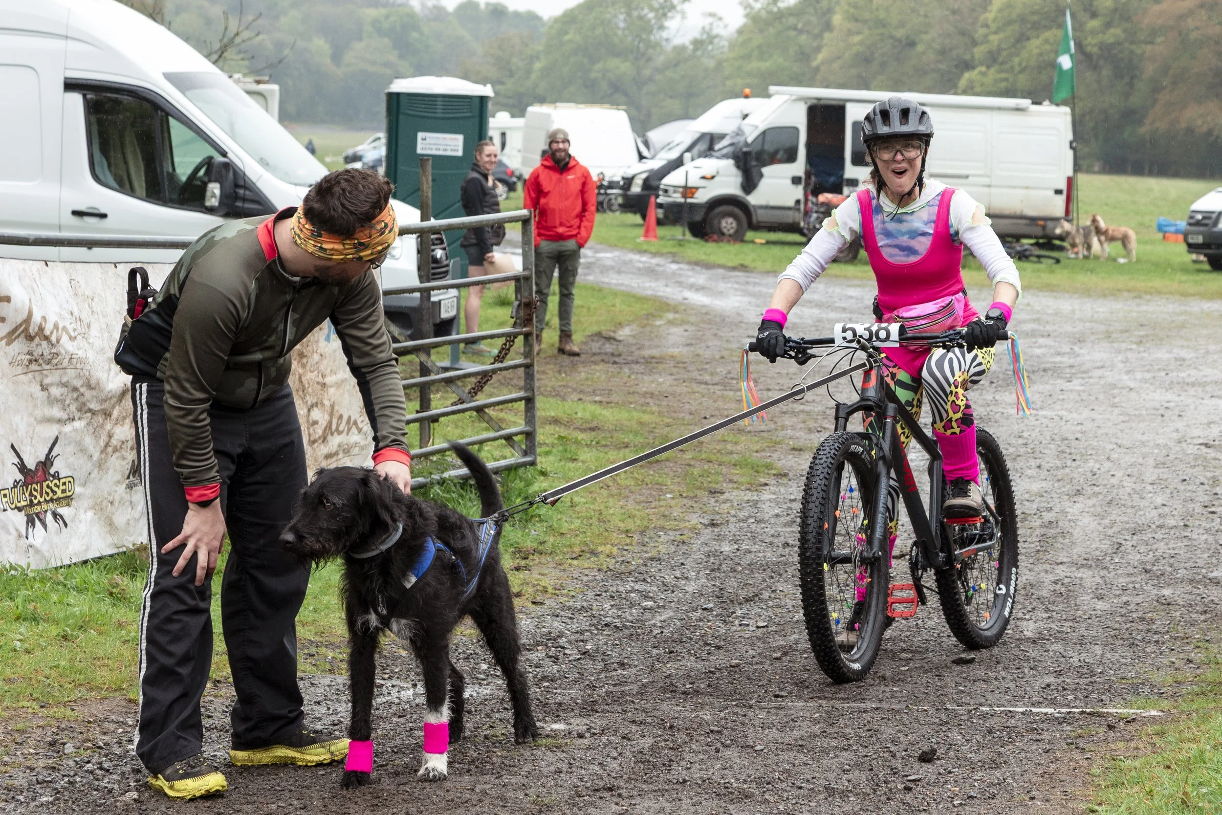 A woman riding a mountain bike with a race bib, smiling, and dressed in colorful, playful clothing and helmet. A man holding a black goat with pink leg warmers stands nearby. Several vehicles and people are in the background at an outdoor event in a 