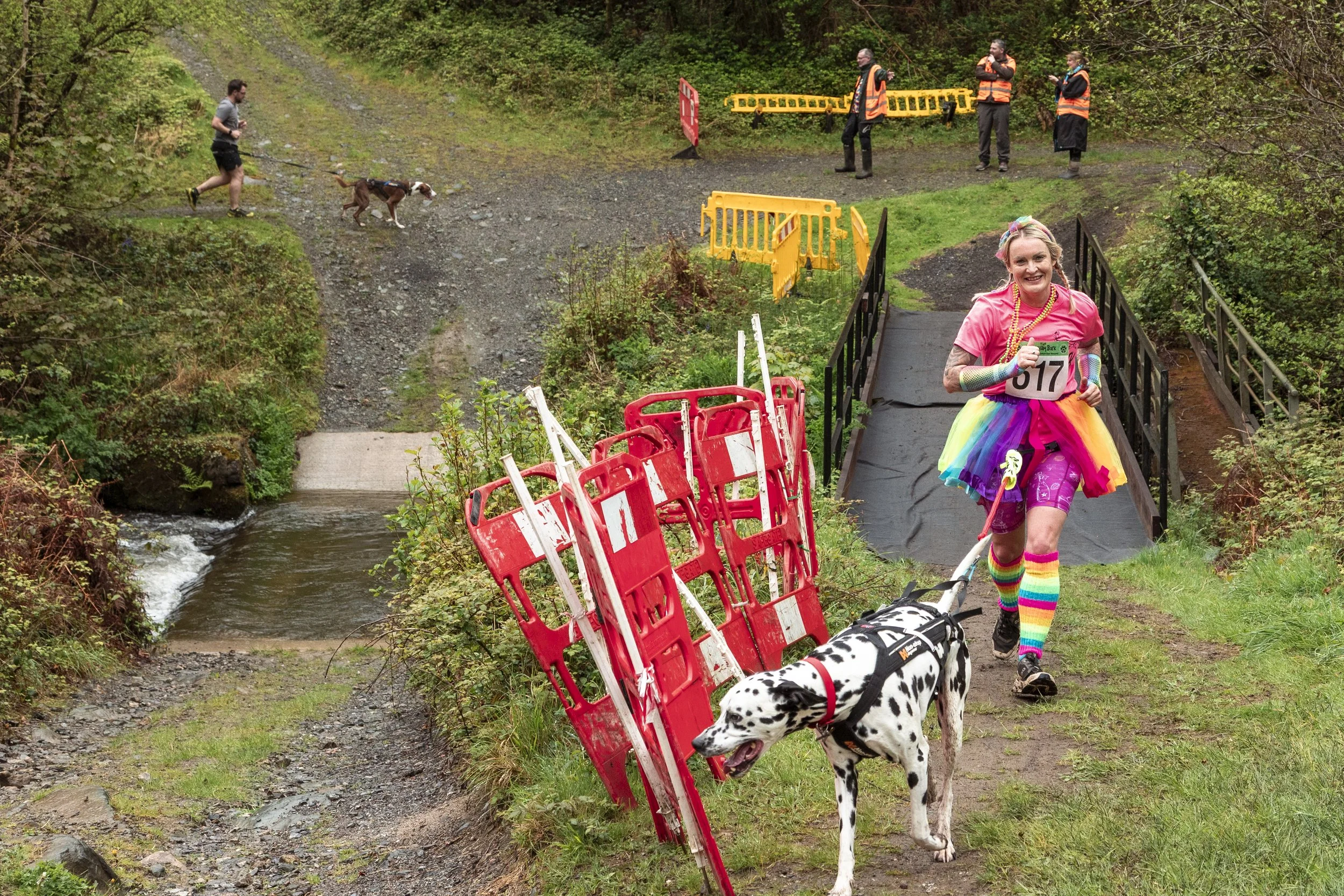 A woman dressed in a colorful rainbow tutu and rainbow striped socks is running a race with a Dalmatian dog on a trail near a small water crossing. In the background, volunteers in orange vests and a person with a dog are near a barricade and yellow 