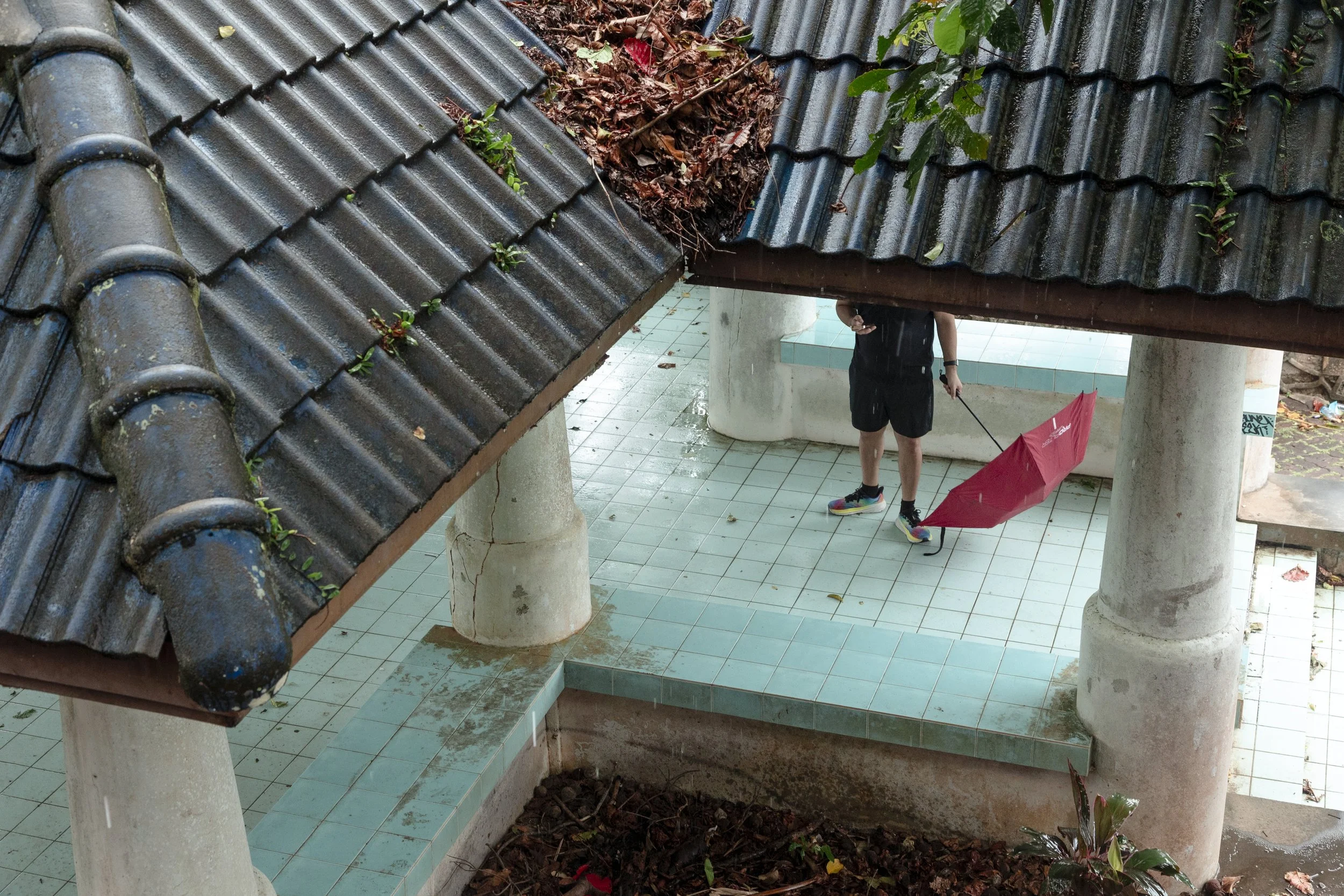 A person holding a red umbrella stands under a tiled roof in an outdoor area with columns and tiled flooring, while rain is falling.