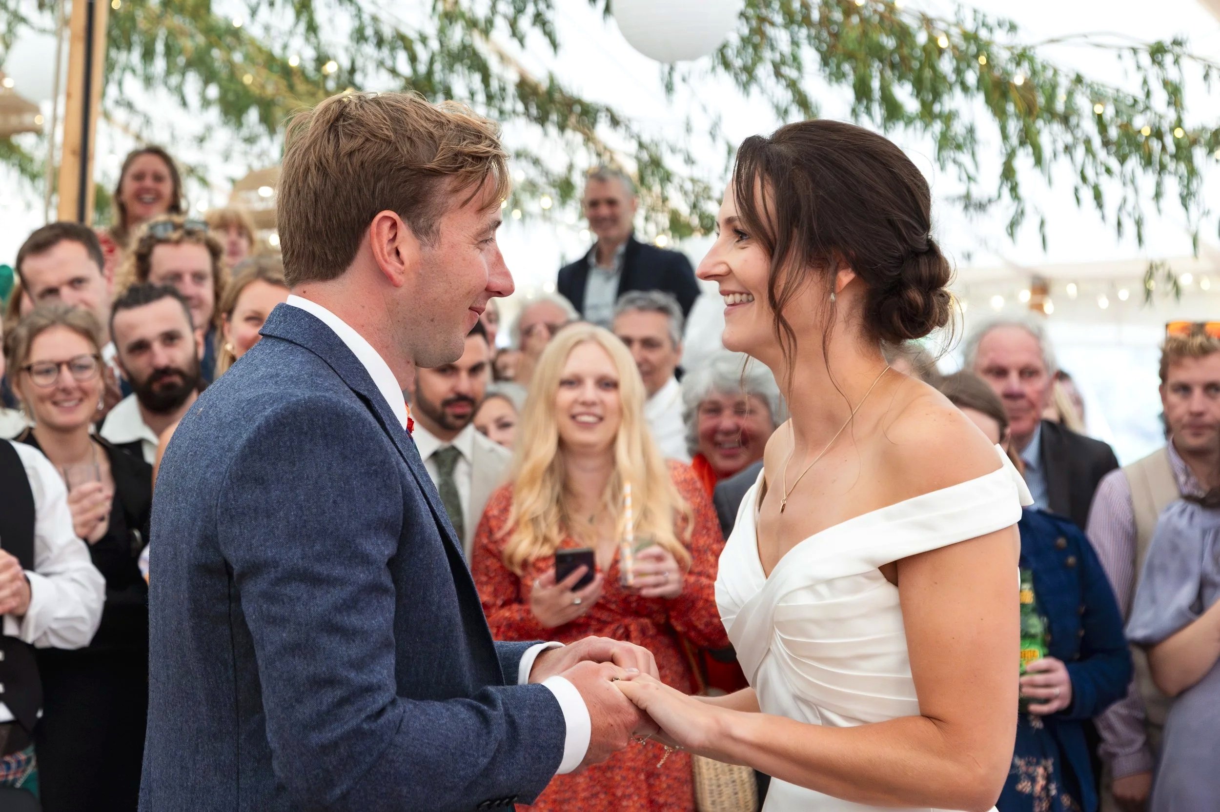 A bride and groom hold hands and smile at each other during their wedding ceremony, surrounded by friends and family in a marquee with string lights and green leaves overhead.