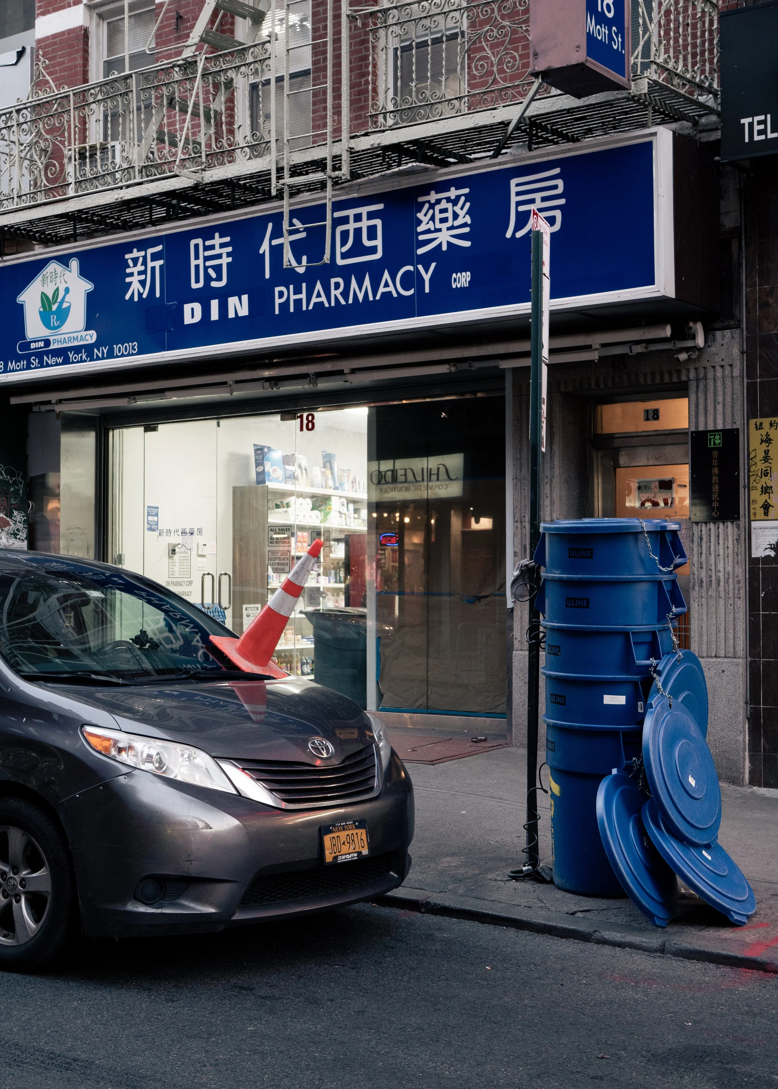 Street view of DIN Pharmacy storefront with blue sign featuring Chinese characters and English name, glass door entrance, a parked black car with a traffic cone on hood, and a blue multi-compartment mail collection box with multiple blue lids stacked