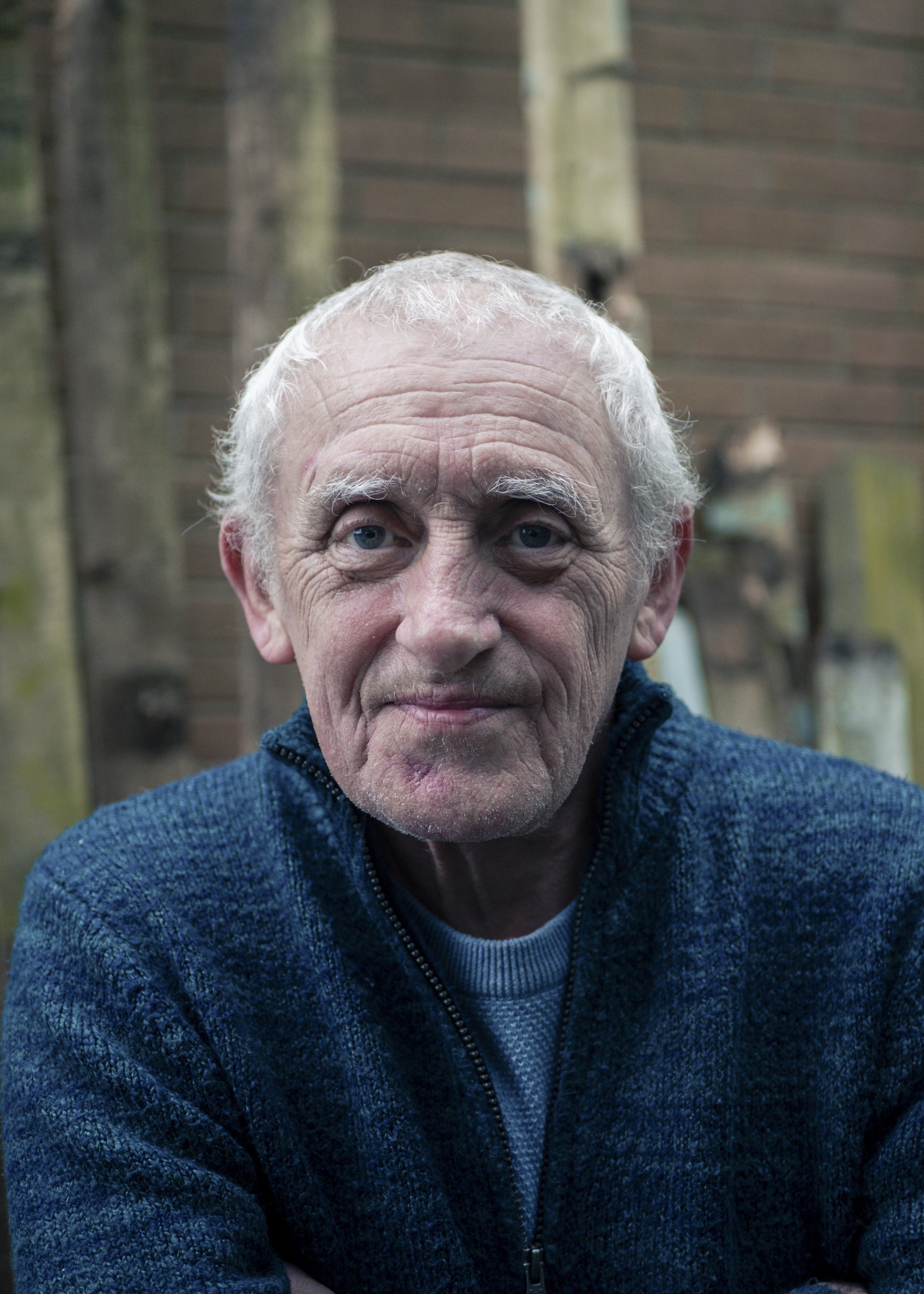 Close-up of an elderly man with white hair and blue eyes, wearing a dark blue sweater, sitting outdoors in front of a wooden background.