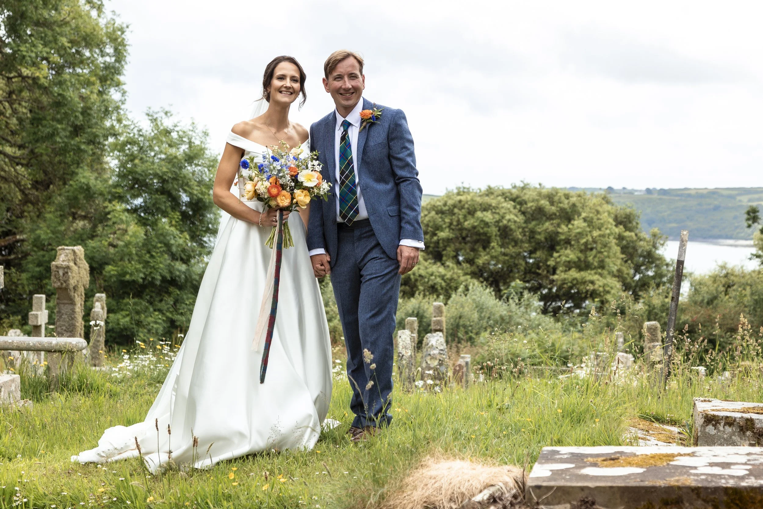 A bride and groom holding hands and smiling in a green outdoor setting with trees and a body of water in the background. The bride is wearing a white wedding dress and holding a colorful bouquet. The groom is wearing a blue suit with a patterned tie.
