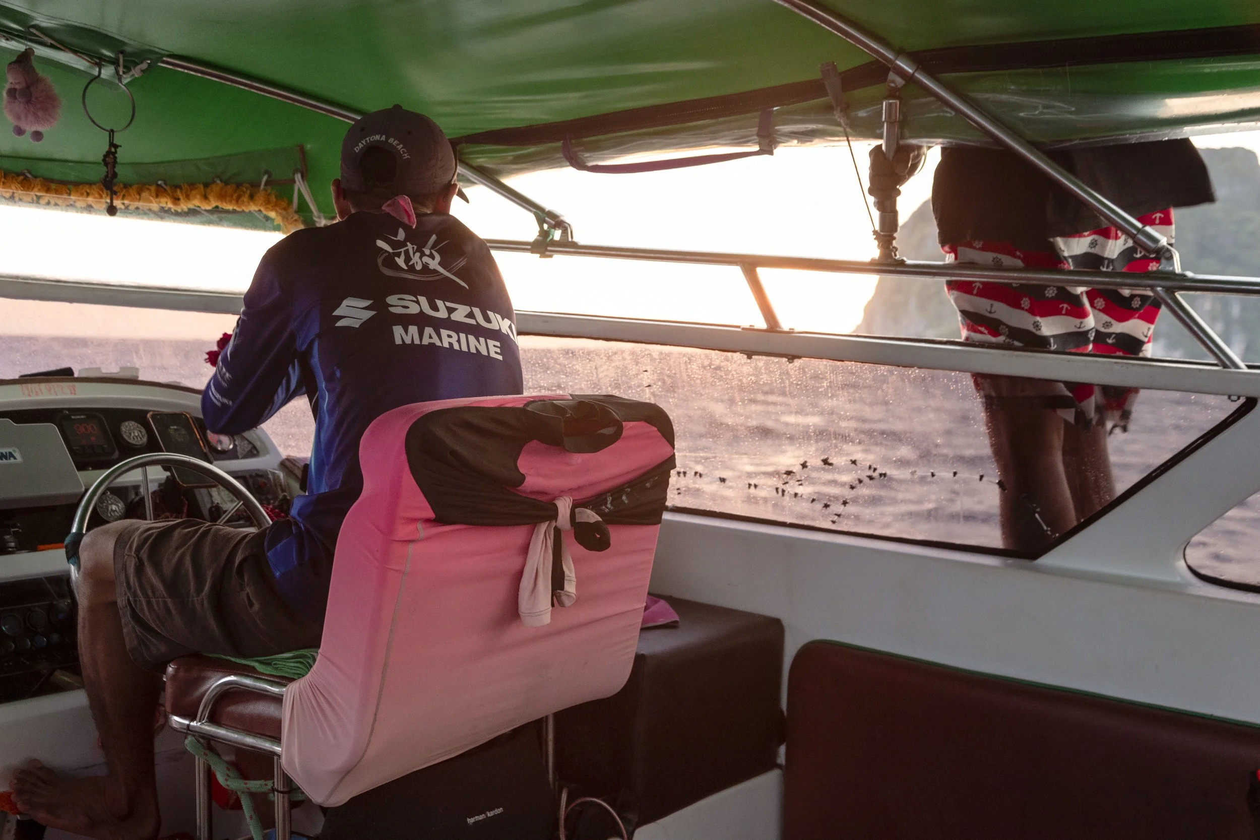 A man on a boat at sunset, wearing a navy blue long-sleeve shirt with Suzuki Marine logo, sitting at the helm, with a pink towel draped over the seat. He is looking out towards the water, and another person standing outside the boat is wearing shorts