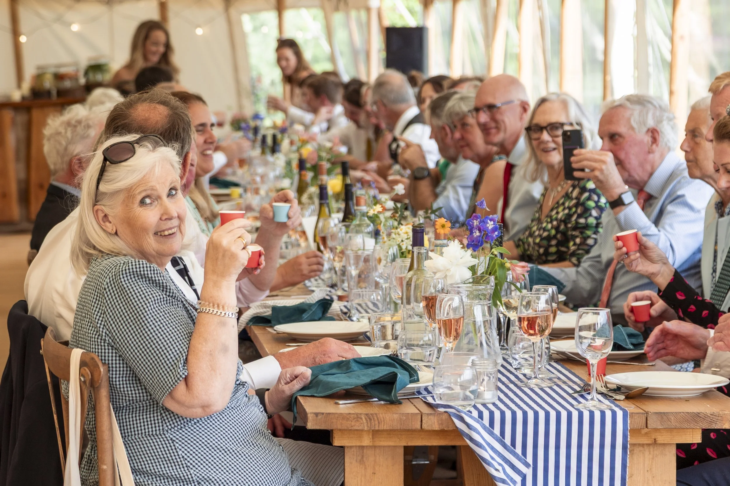 A large group of mostly elderly people sitting at a long dining table inside a bright, wooden-framed venue, celebrating with drinks and smiling, with flowers and bottles on the table.