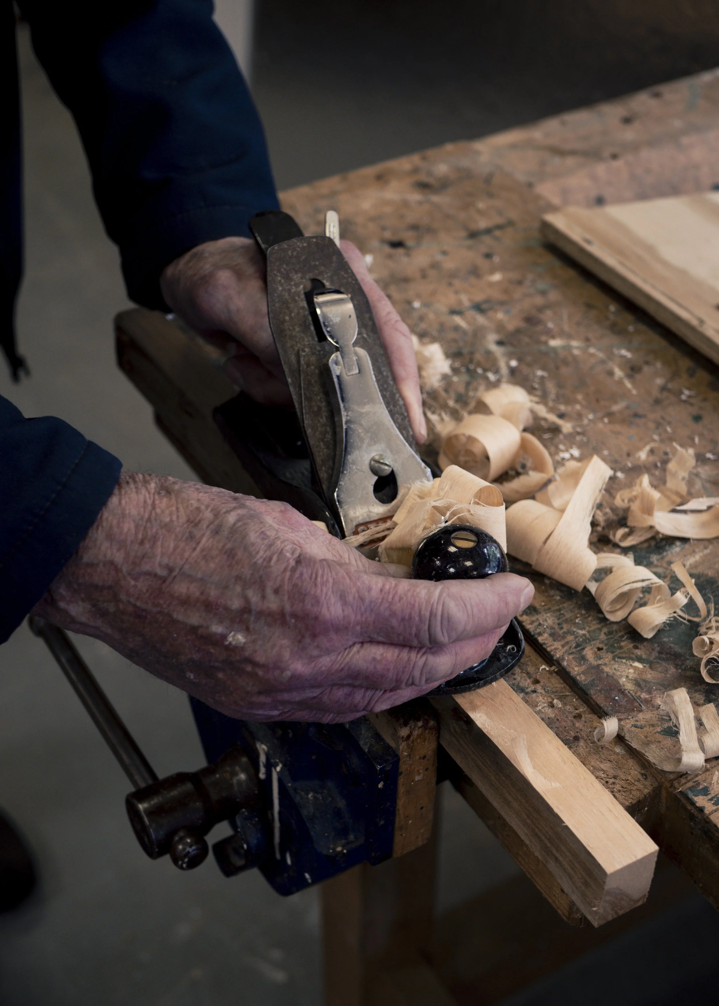 A person with aged hands using a hand plane to shave wood on a workbench with wood shavings around.