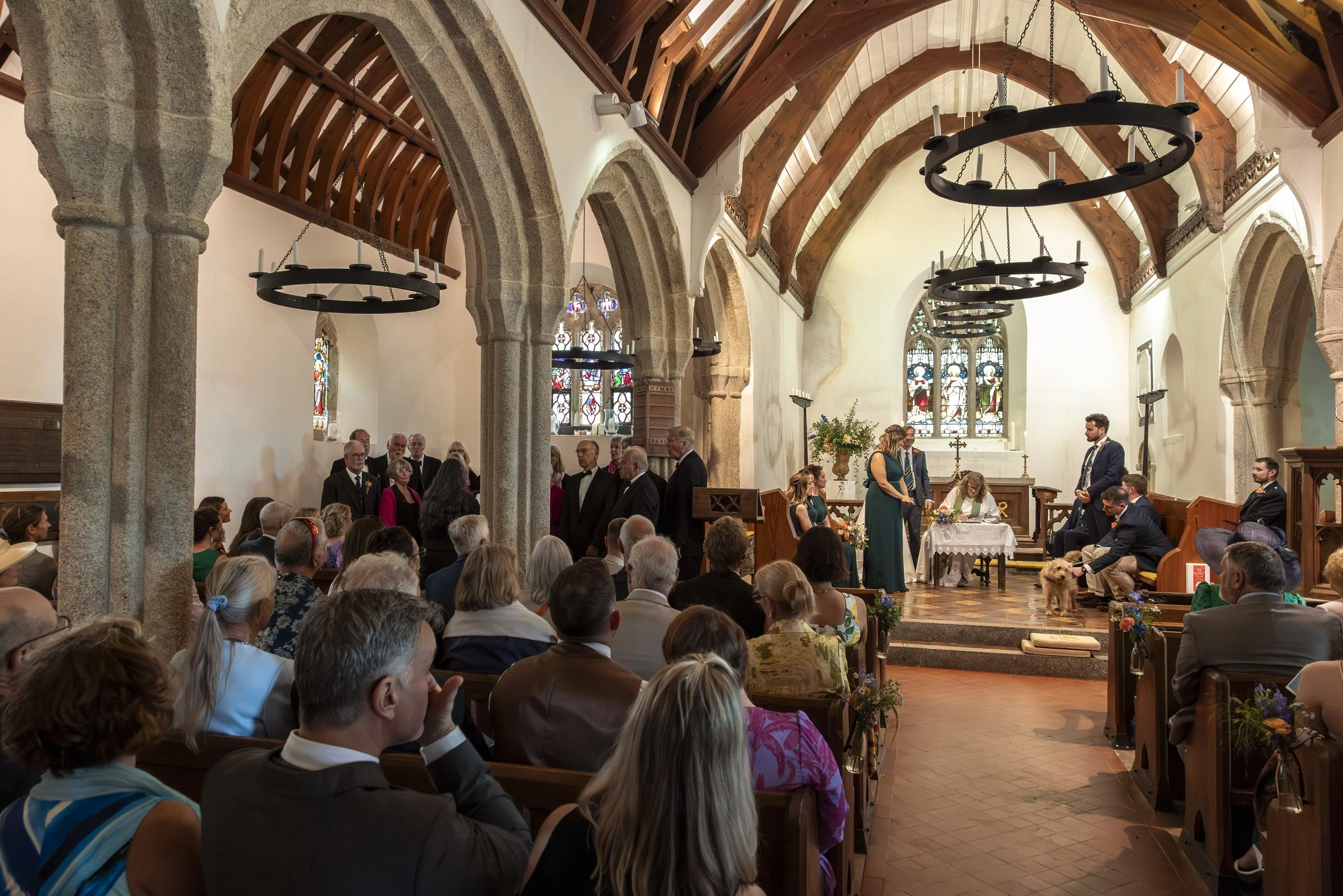 A wedding ceremony inside a church with wooden beams, stained glass windows, and an altar. Guests seated in pews watch the couple exchange vows, with the officiant and bridal party present.