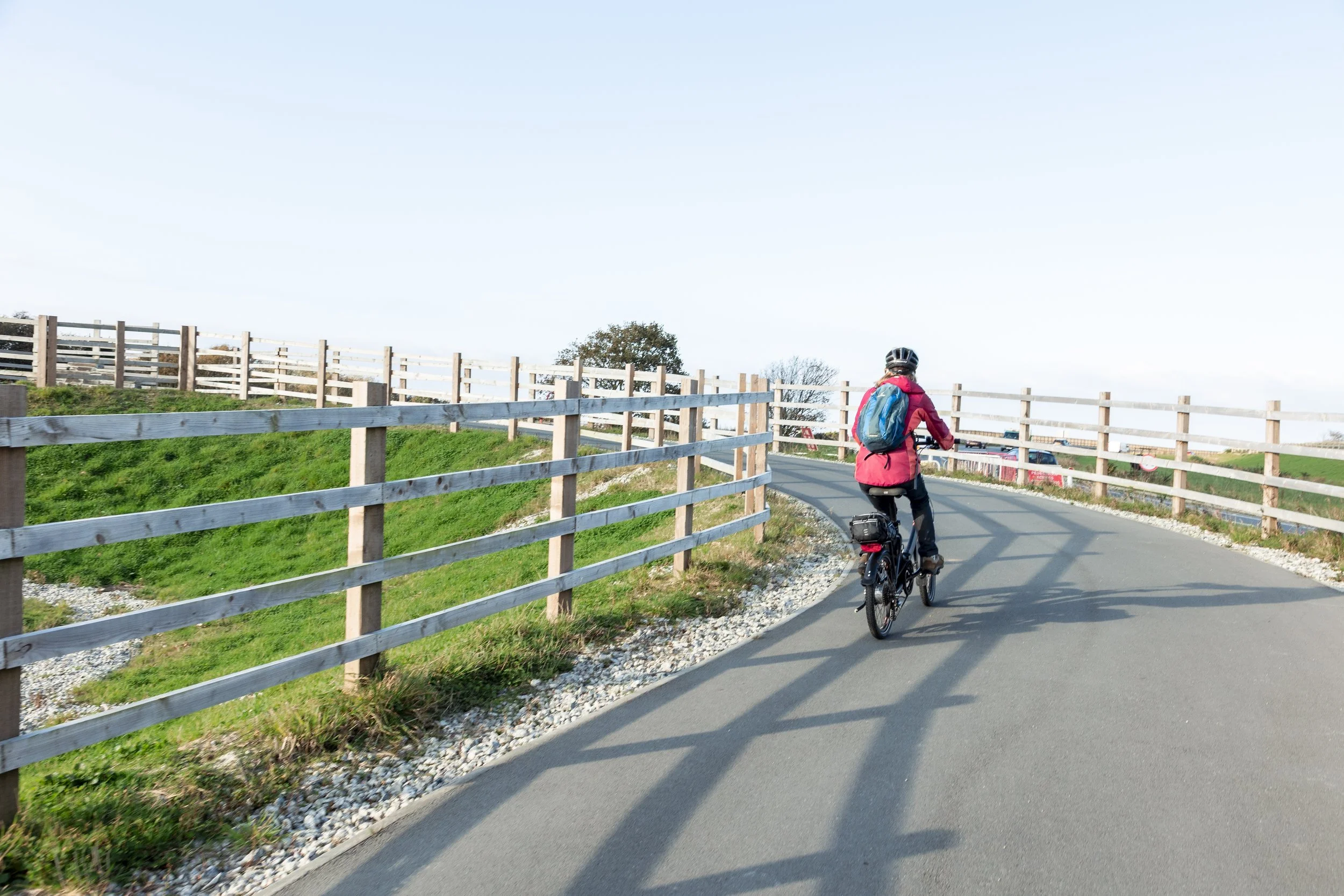 A person biking on a paved path, wearing a helmet, red jacket, and carrying a backpack, with wooden fences lining both sides of the path in a rural setting.