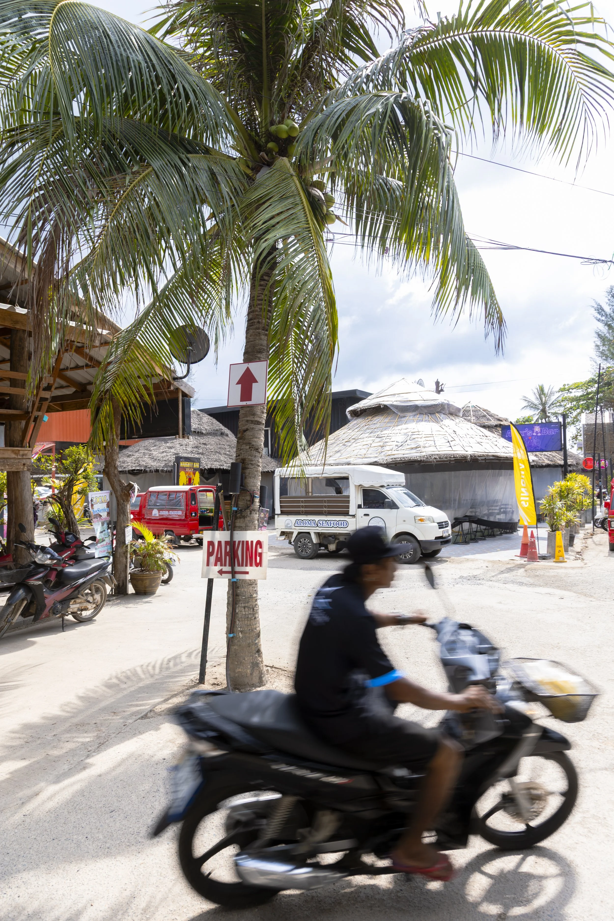 A tropical street scene featuring a tall palm tree, a motorcycle rider, parked motorcycles, a white van, thatched-roof buildings, and signs for parking and an arrow pointing straight ahead, with partly cloudy sky.