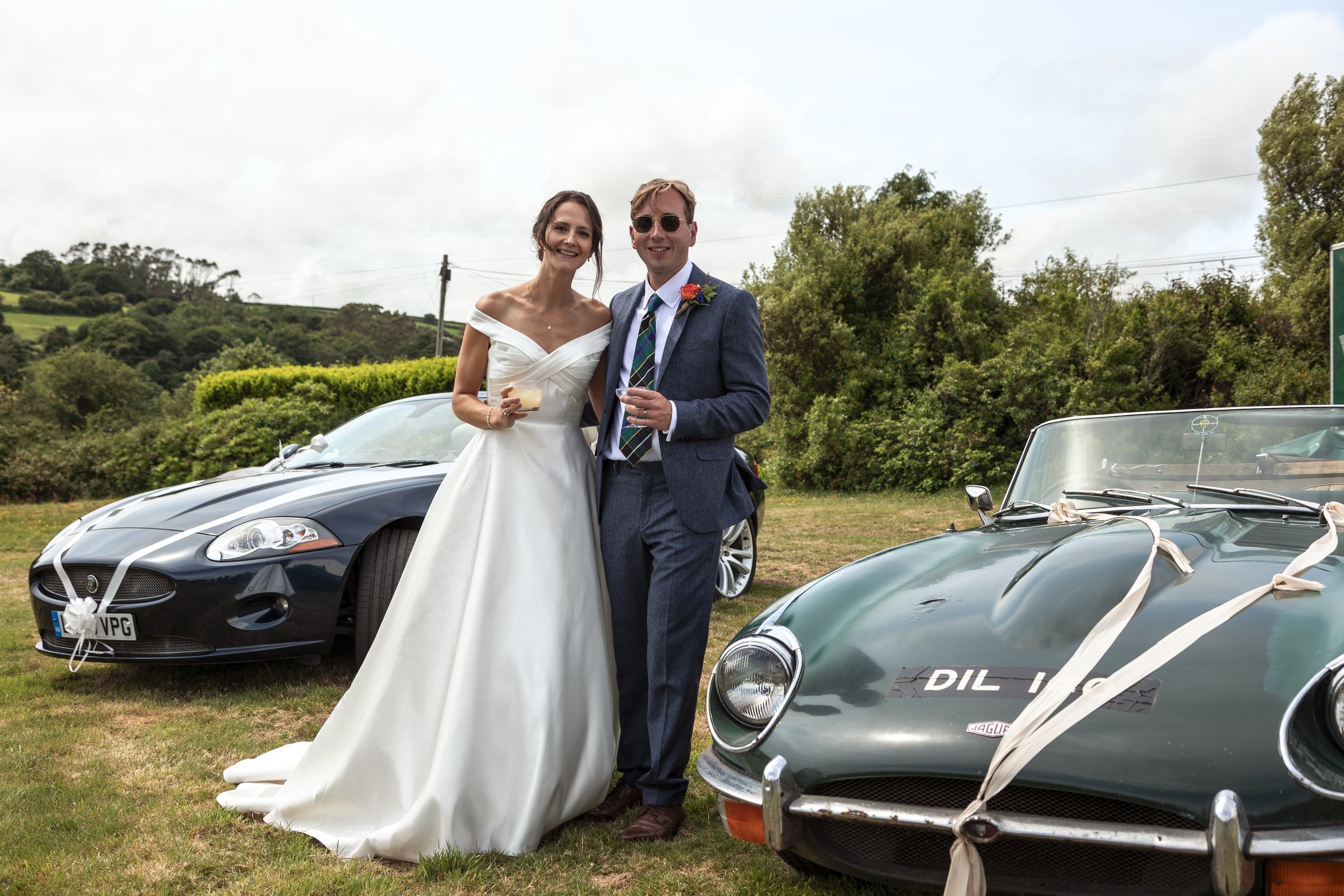 A bride and groom at a wedding standing outdoors between two vintage cars, with greenery and hills in the background. The bride is in a white wedding dress holding a drink, and the groom is in a suit with sunglasses, also holding a drink.