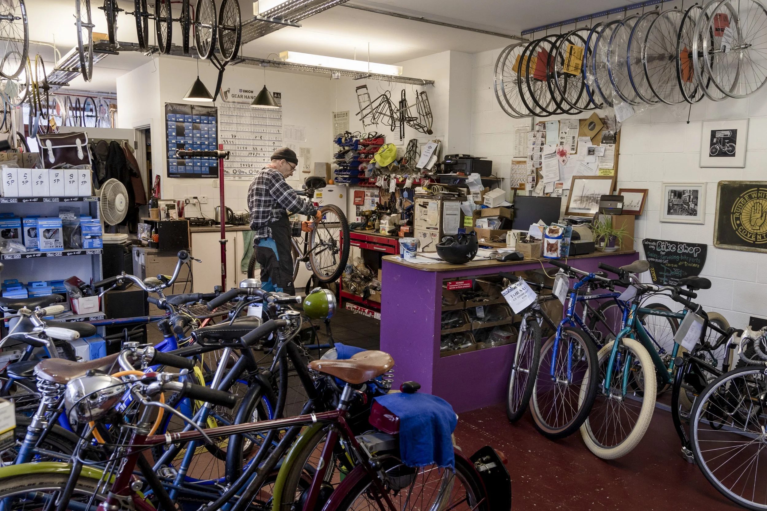 Bike shop with multiple bicycles both on the floor and hanging from the ceiling, a mechanic working at a workbench, tools, and bike parts organized on the walls.