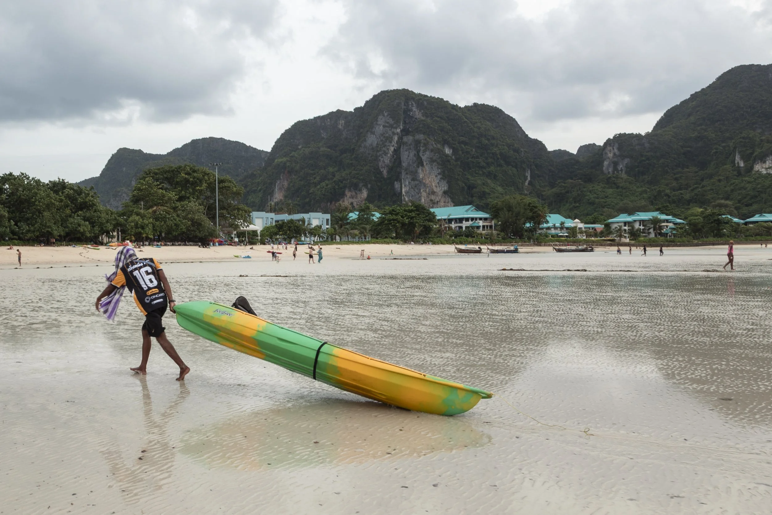 Person walking on sandy beach carrying a green and yellow surfboard, overcast sky, distant mountains, beach huts, and people in the background.