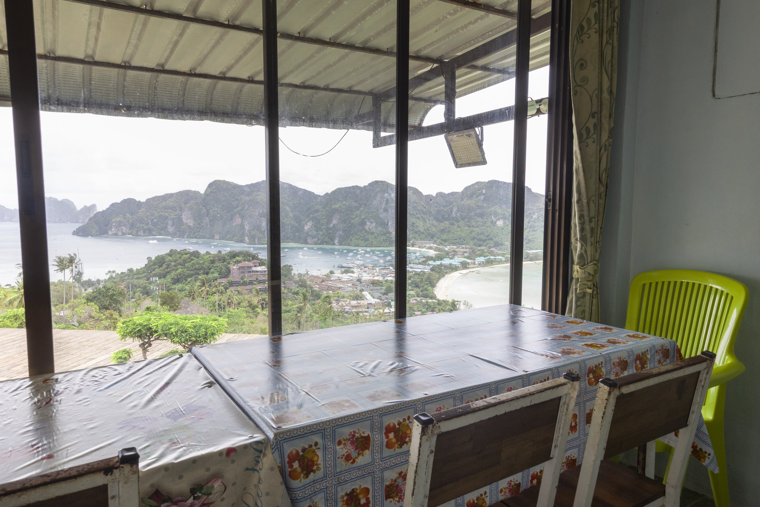 Dining table with floral tablecloth near large glass windows showing a coastal landscape with mountains, water, and boats outside.